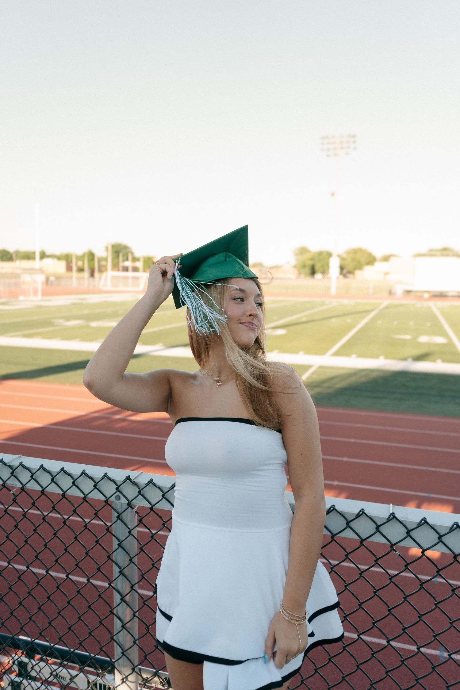 Young woman in white dress poses with green graduation cap at athletic field.