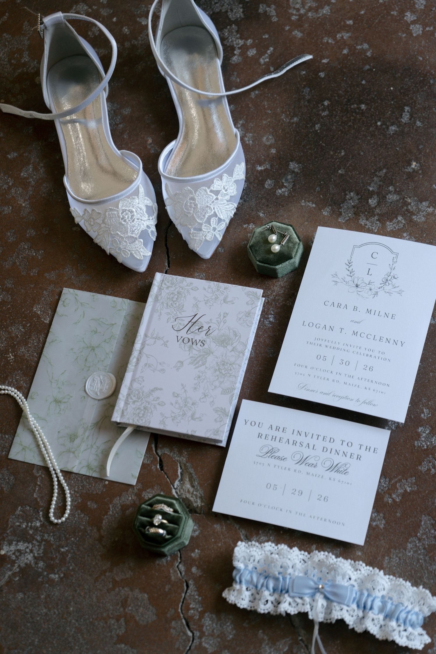 Elegant wedding flat lay featuring lace bridal shoes, vow books, pearl necklace, ring box, garter, and invitation suite.