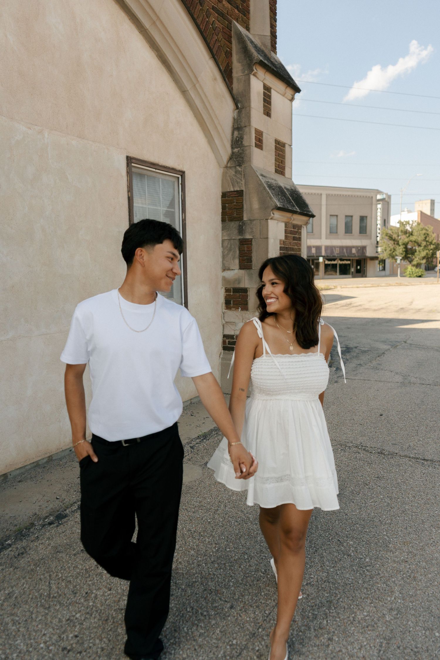 Young couple in matching white outfits holding hands and smiling while strolling past a building.