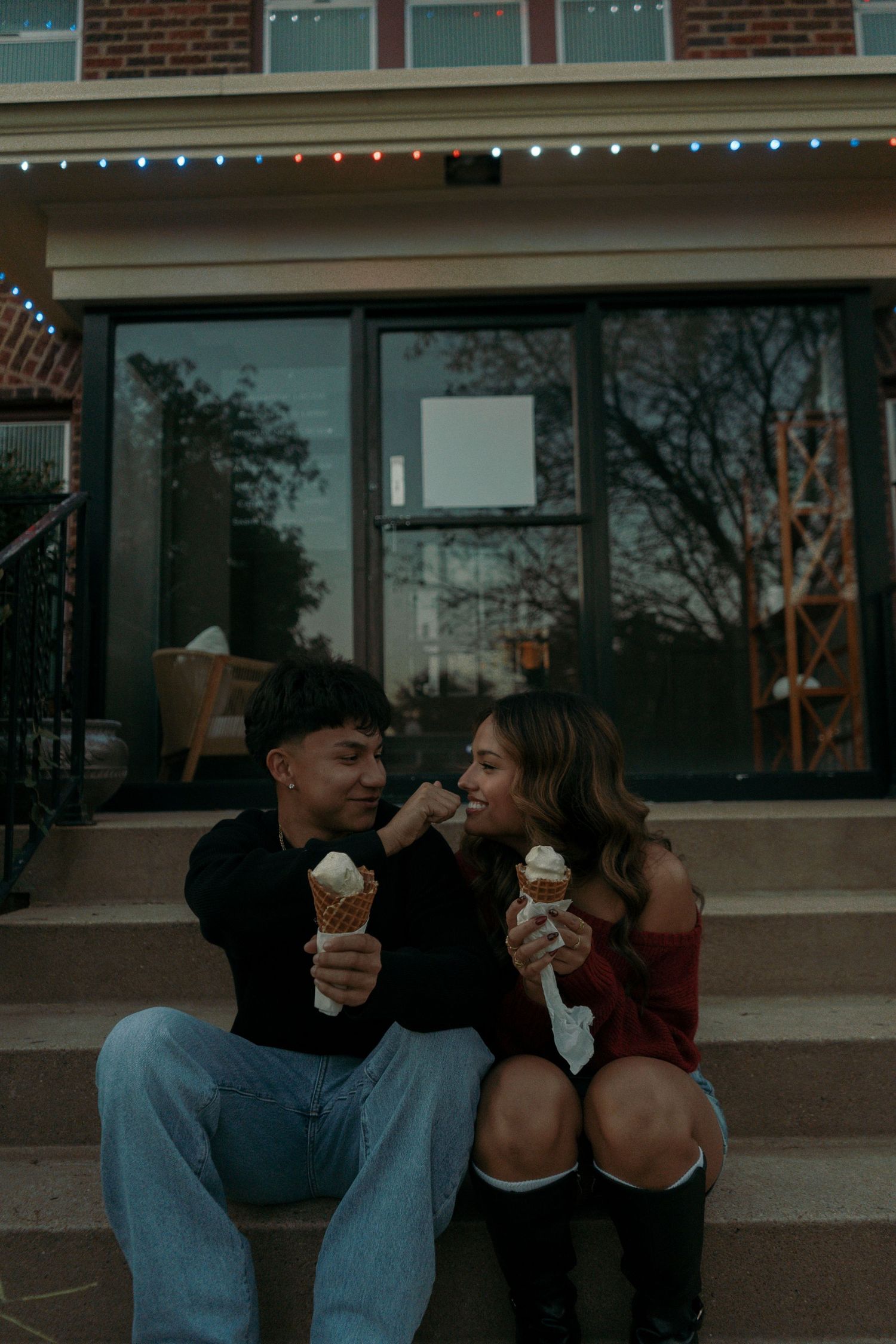 Happy couple sitting on steps sharing ice cream cones outside a building with string lights at dusk.