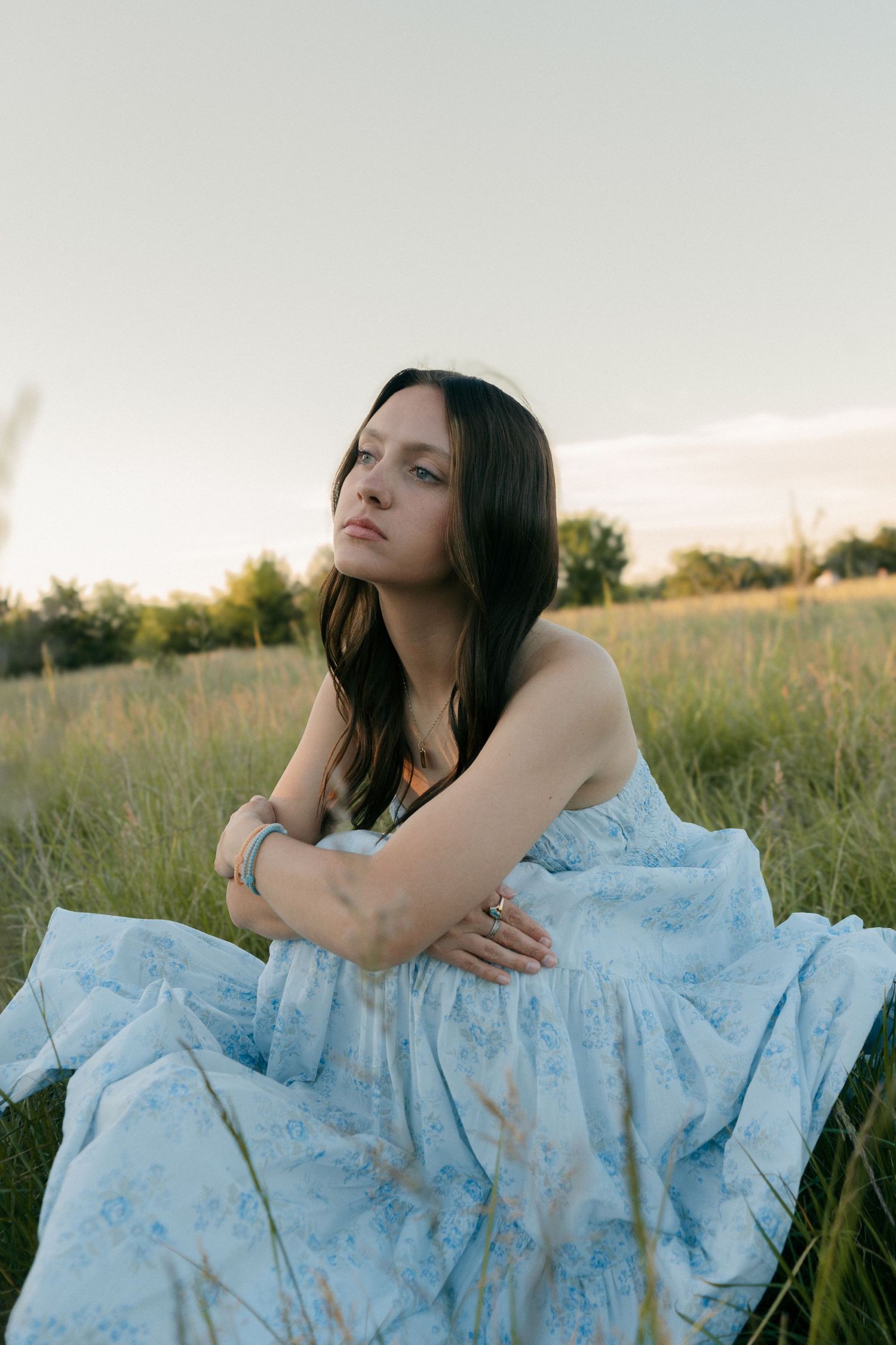 Brunette woman in blue floral dress sitting in grassy field at golden hour with arms crossed.
