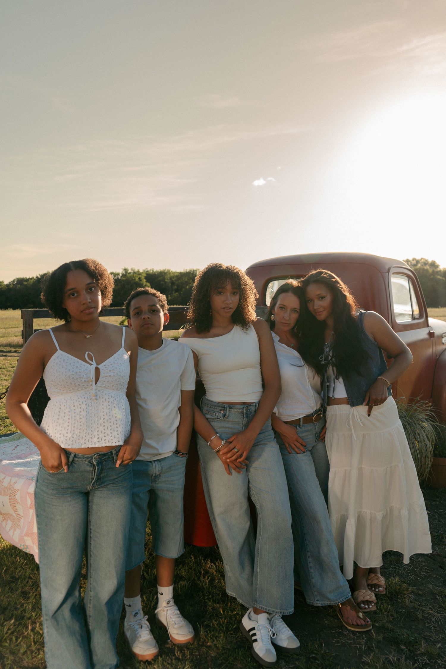 Five young women pose in jeans and white tops beside a vintage red truck in a sunlit field.