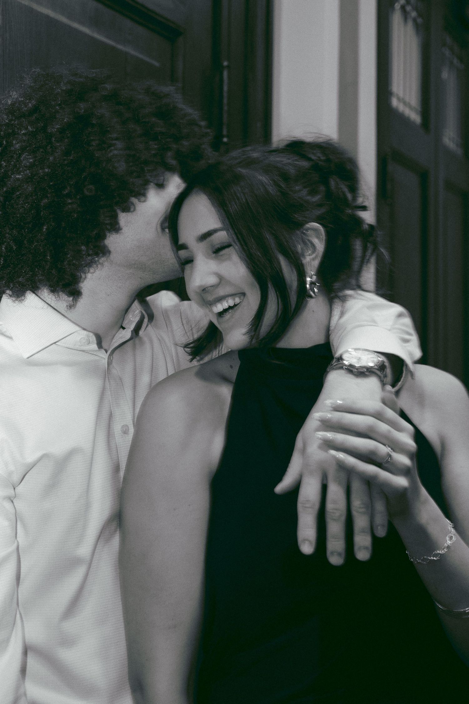Couple sharing a joyful embrace, man whispering to smiling woman in black dress, black and white photo.