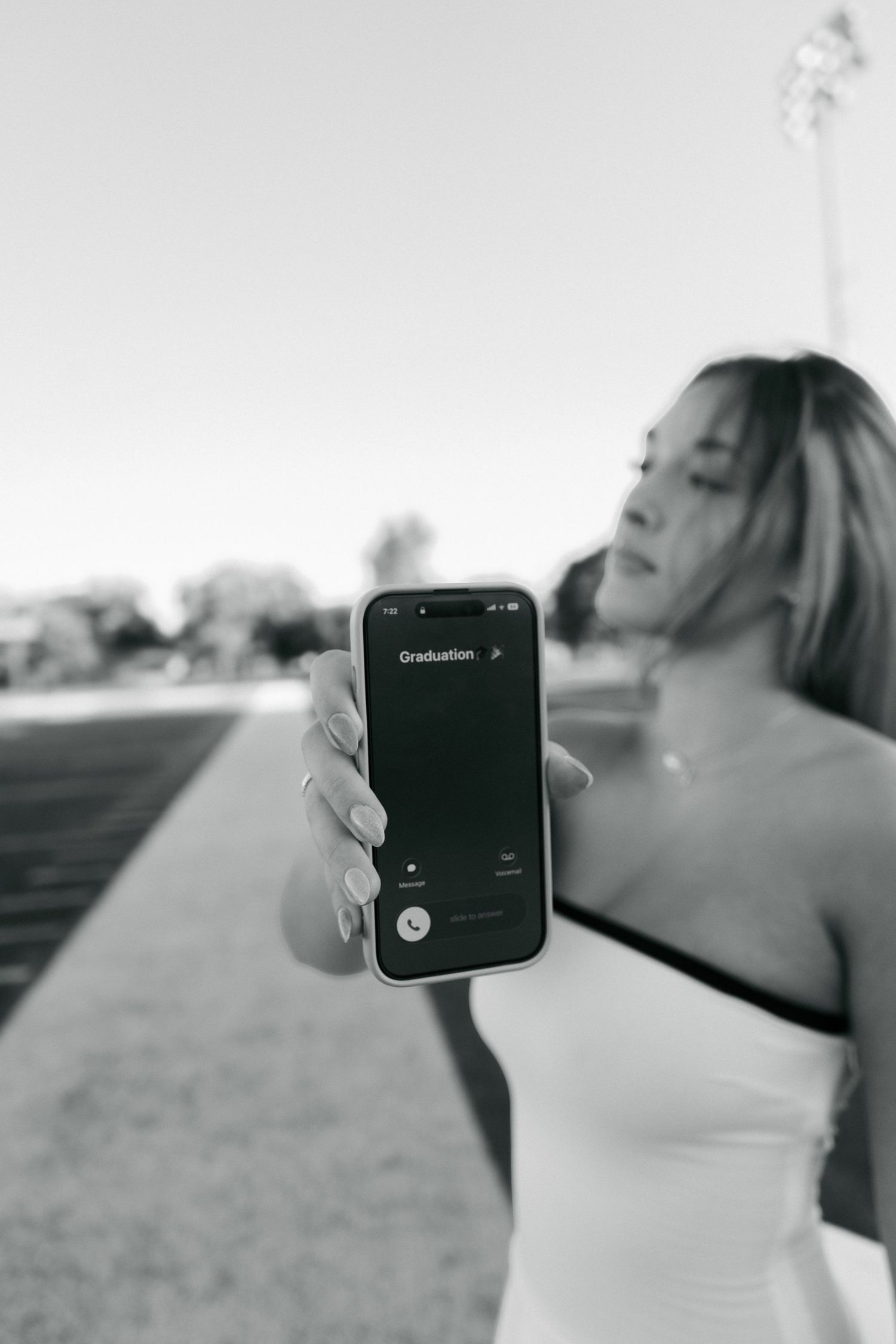 Black and white photo of young woman holding iPhone showing Graduation screen on athletic track.