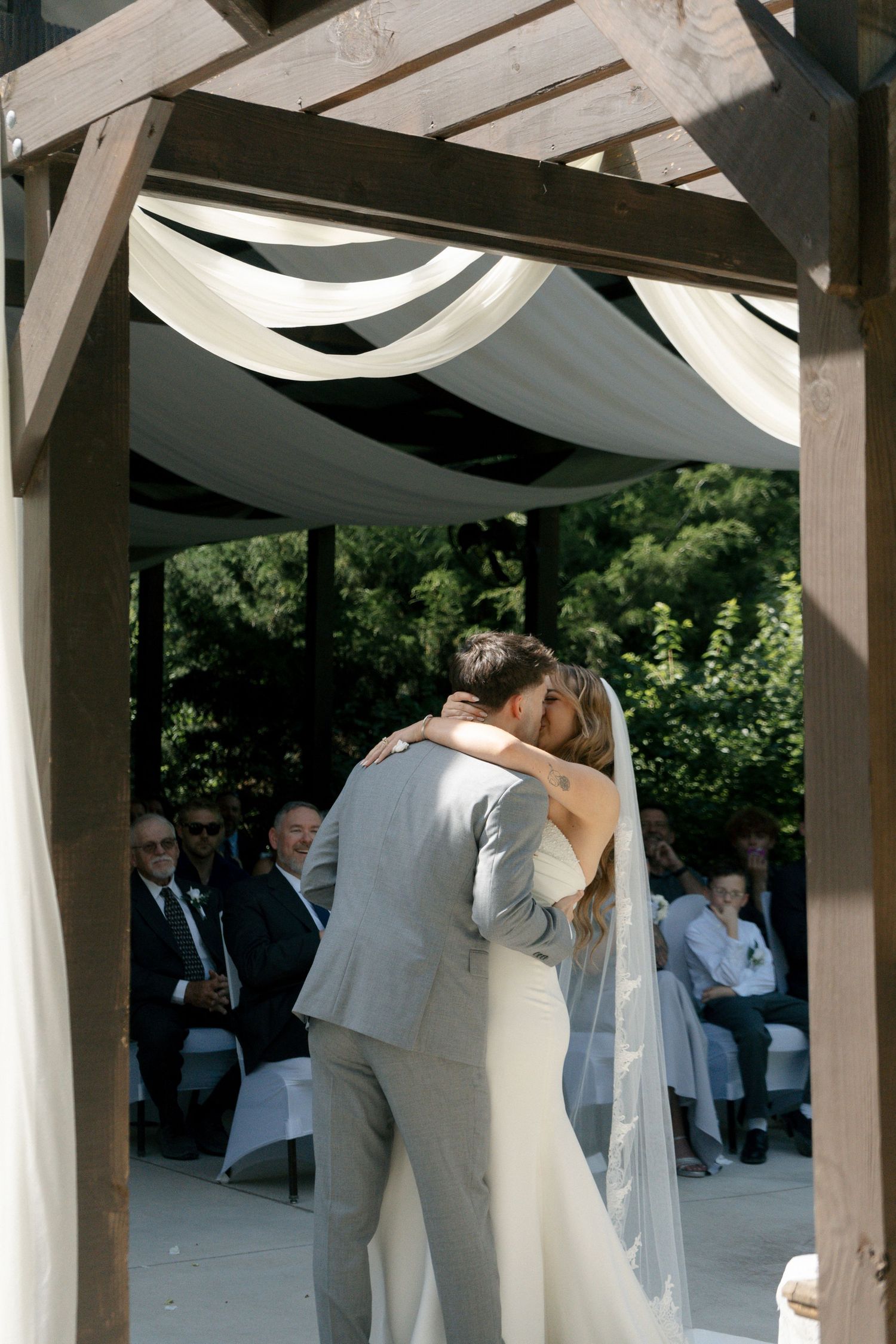 Bride and groom share their first kiss under a draped wooden arch at an outdoor wedding ceremony with guests seated behind.
