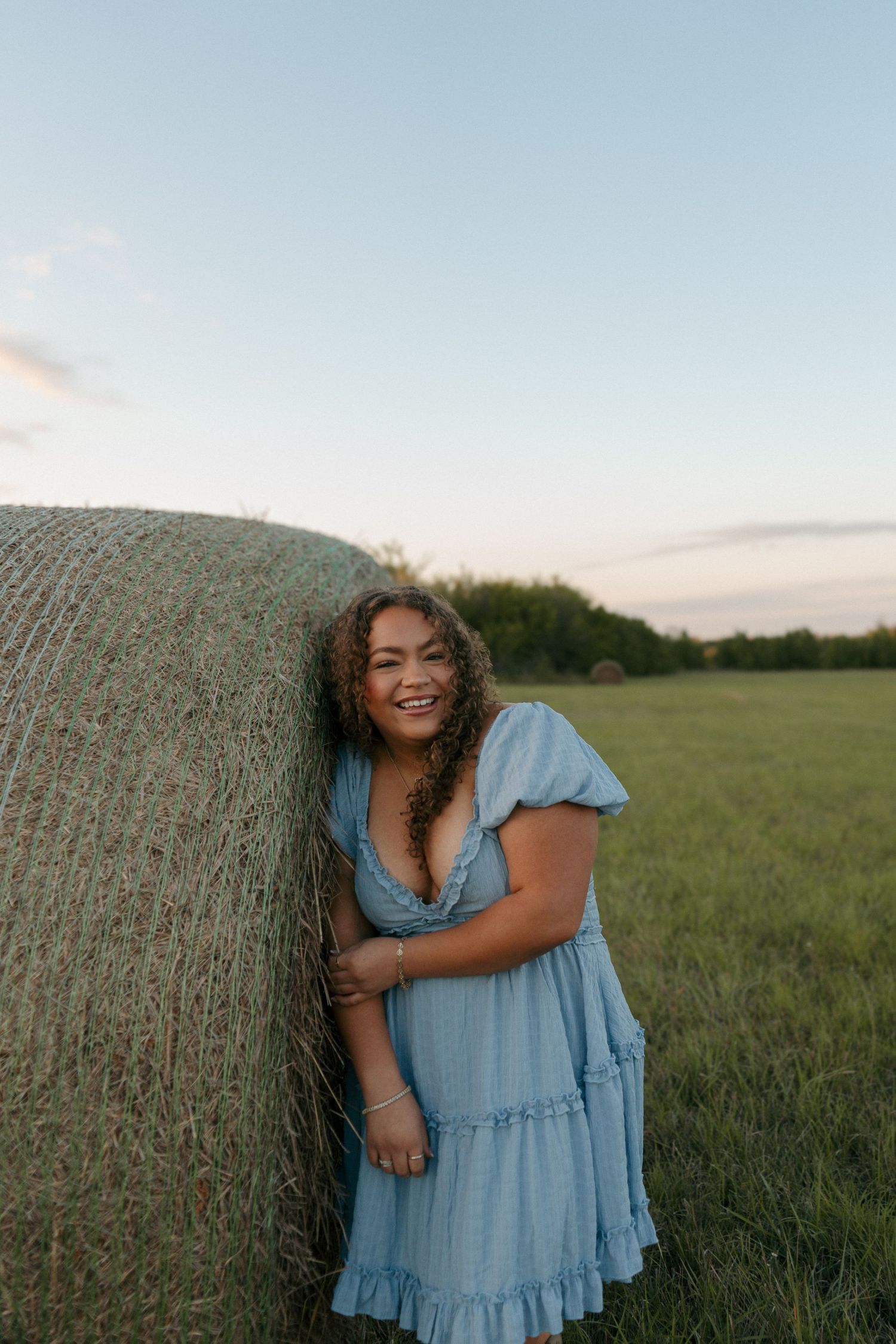 Smiling young woman in light blue dress leaning against a hay bale in a green field at sunset.