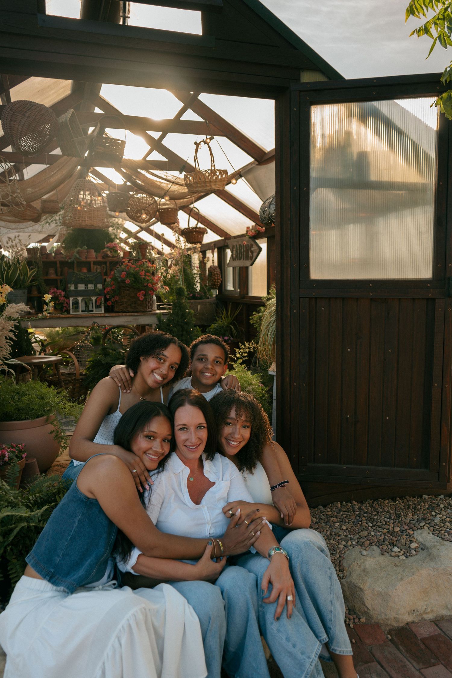 Five women smiling and hugging together outside a sunlit greenhouse surrounded by lush plants.