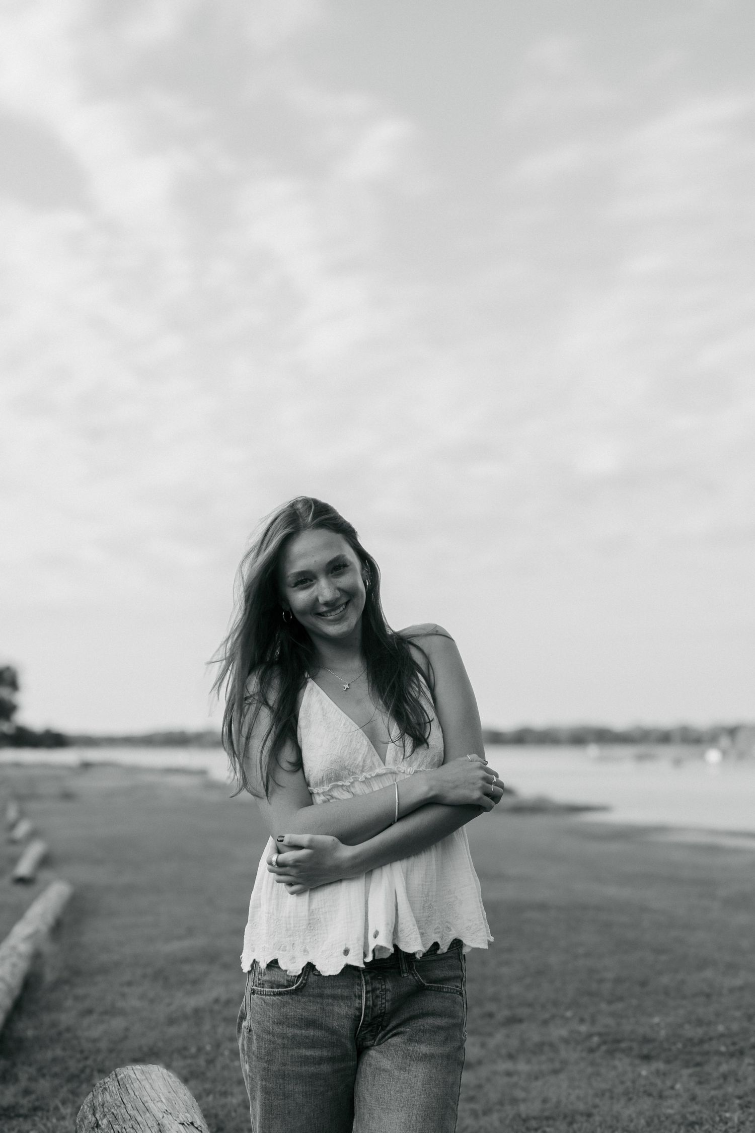 Smiling young woman with flowing hair in white top and jeans posing near waterfront in black and white.