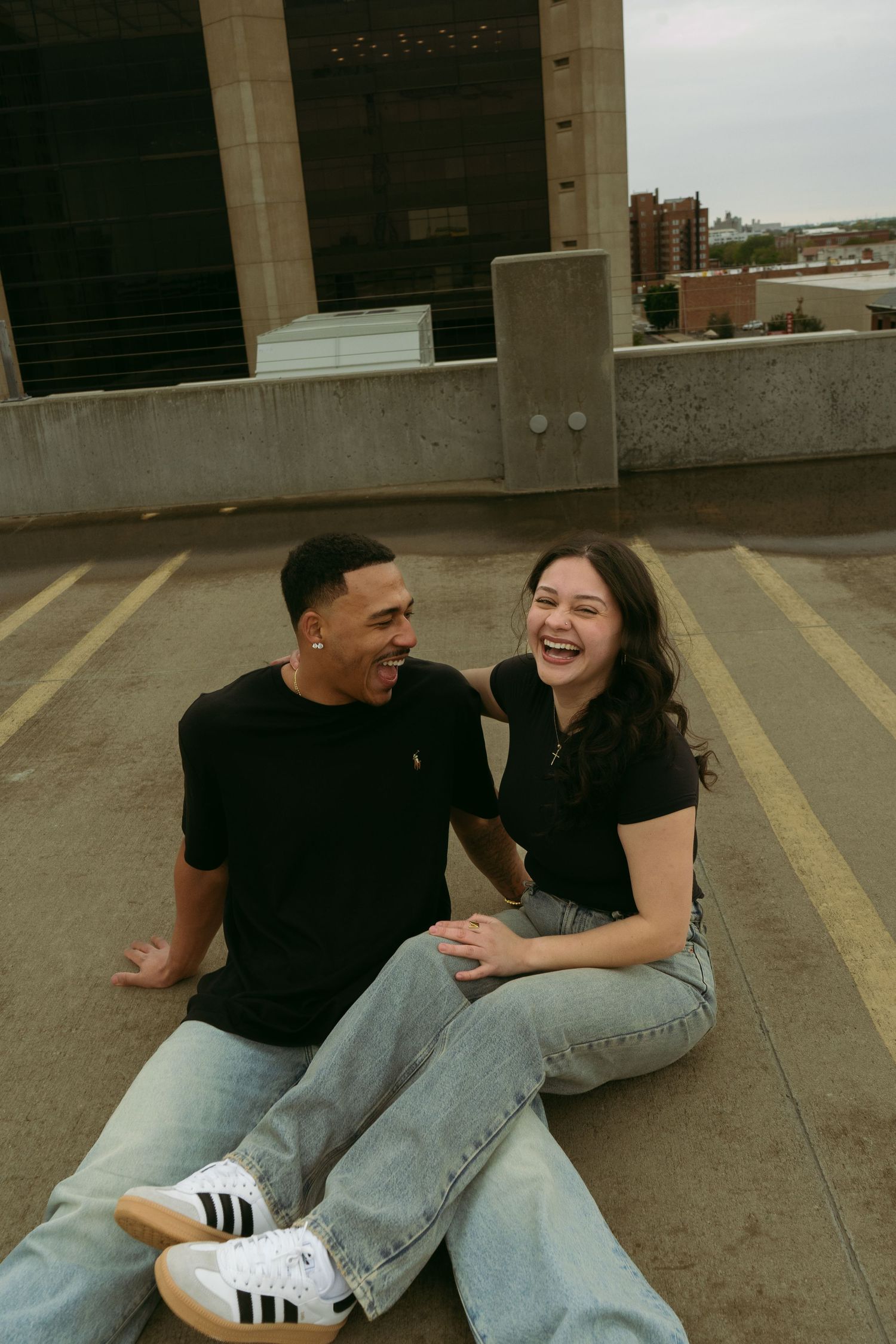 Laughing couple sitting together on a rooftop parking garage, enjoying a candid joyful moment.