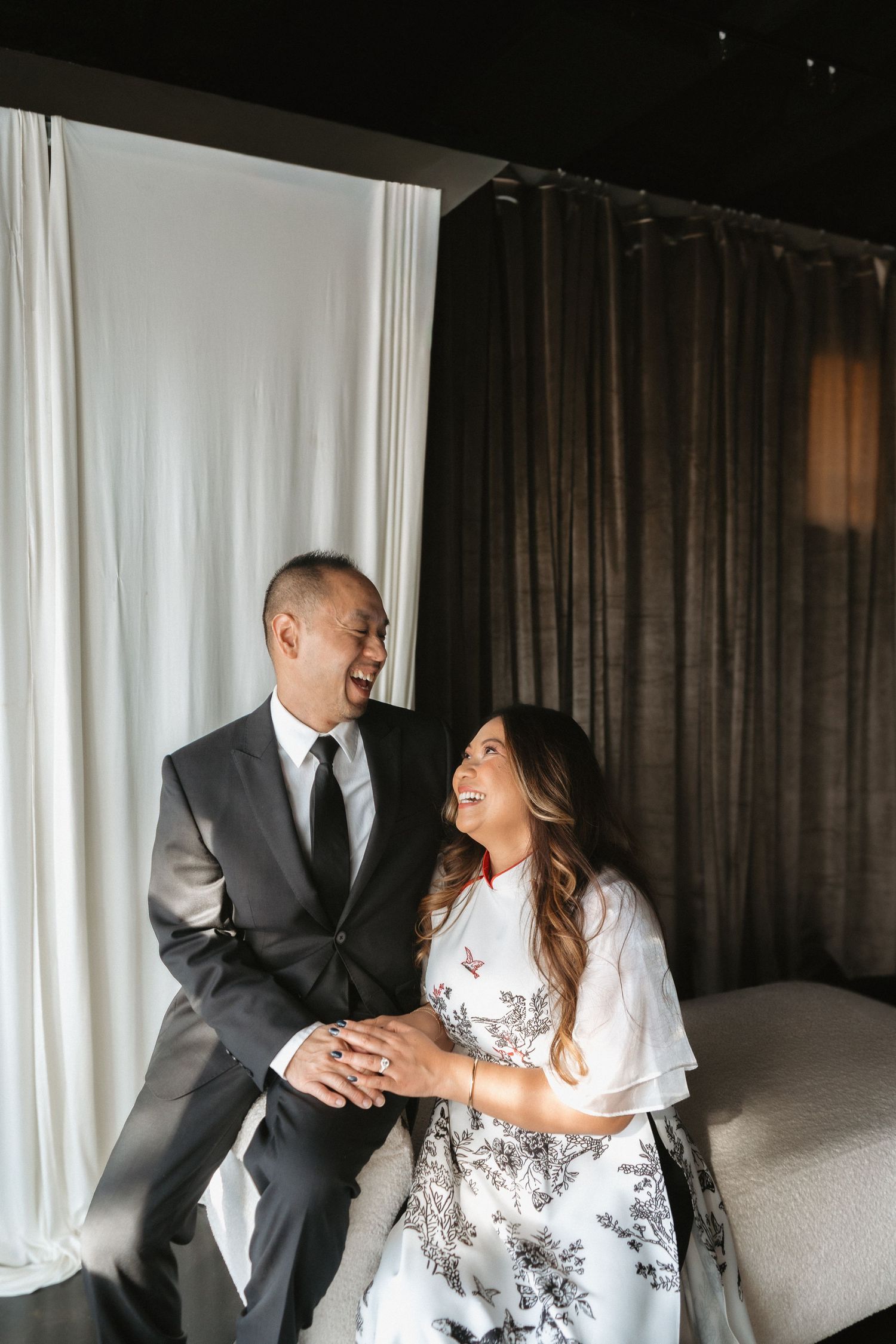 A laughing couple in wedding attire sits together on a couch in a modern, elegant venue.