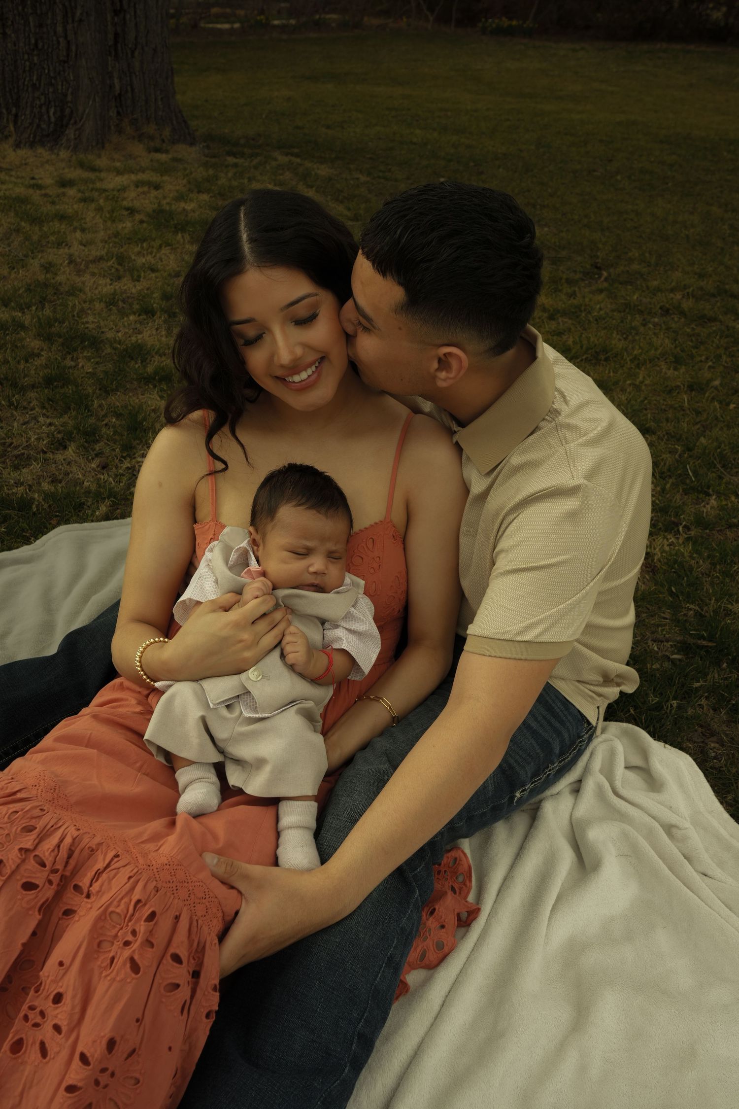 Smiling mother holds newborn while father kisses her cheek as family sits on blanket outdoors.