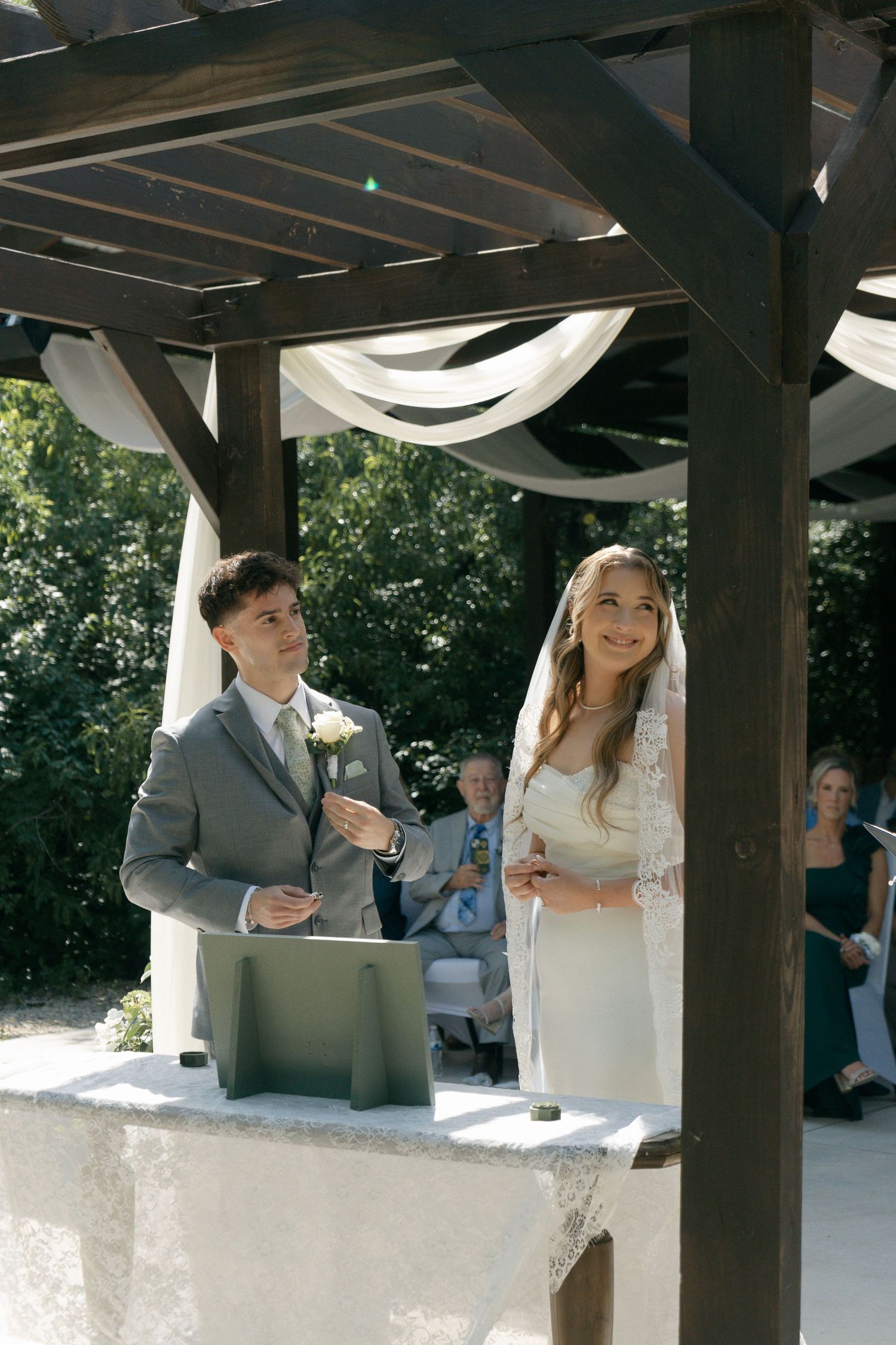 Bride and groom exchange vows under a wooden pergola decorated with white draping at an outdoor wedding ceremony.