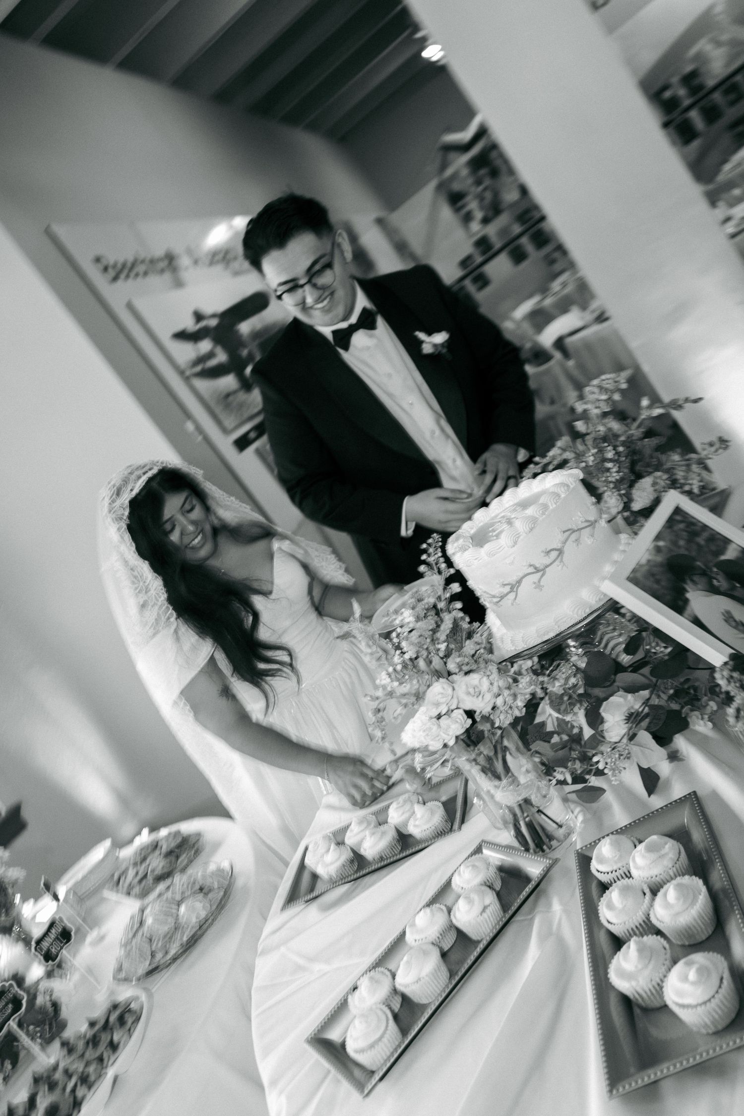 Black and white photo of a bride and groom cutting their wedding cake surrounded by cupcakes at their reception.