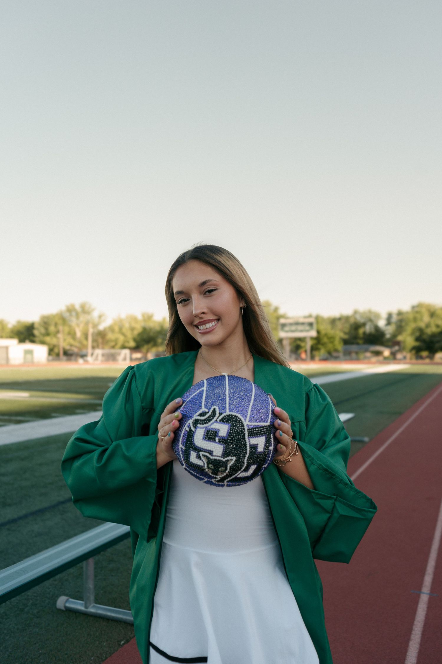 Graduate in green gown holds decorated volleyball on athletic track field.
