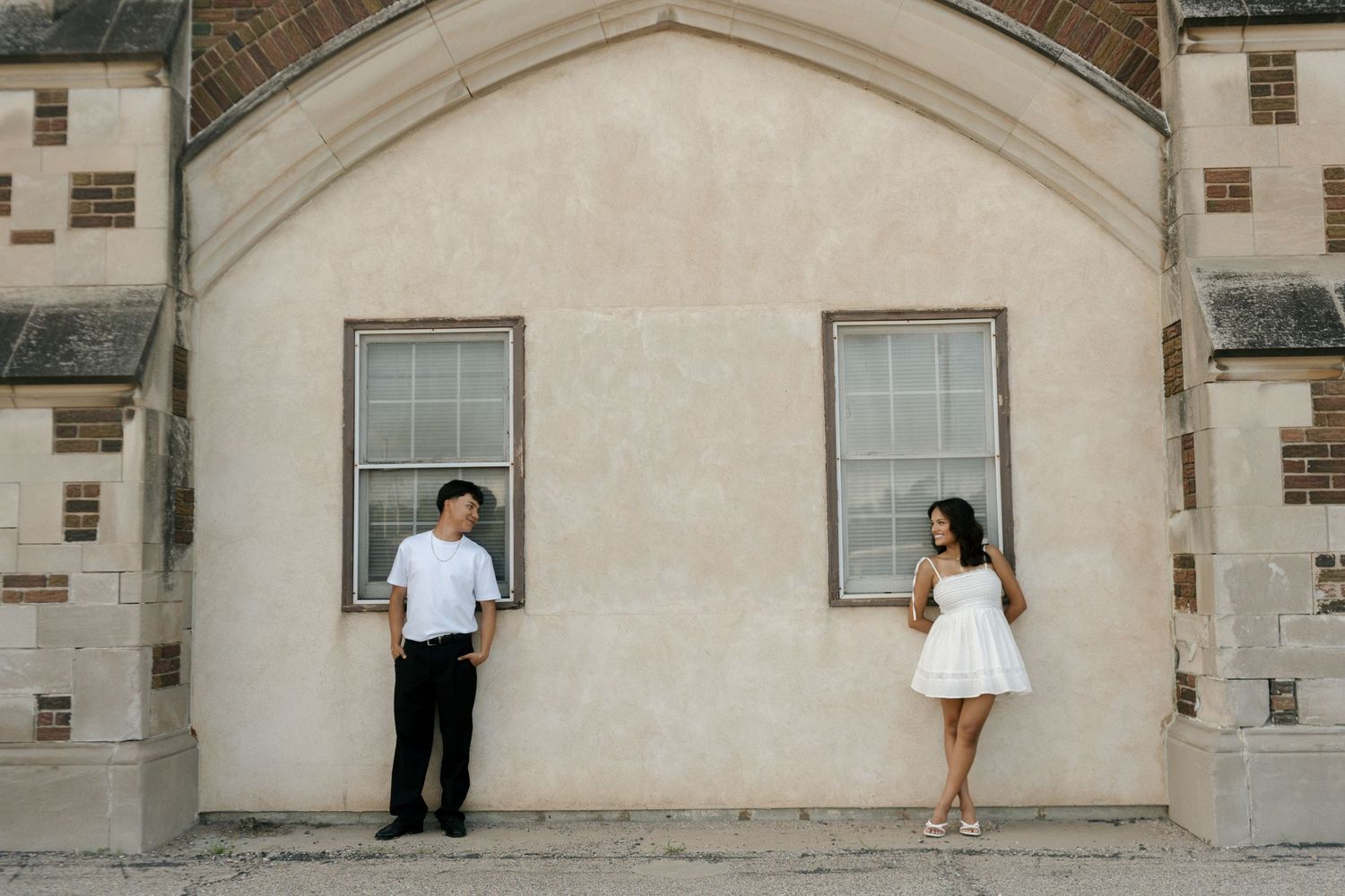 Couple in white outfits standing apart against a beige stone building wall during an outdoor engagement photo session.