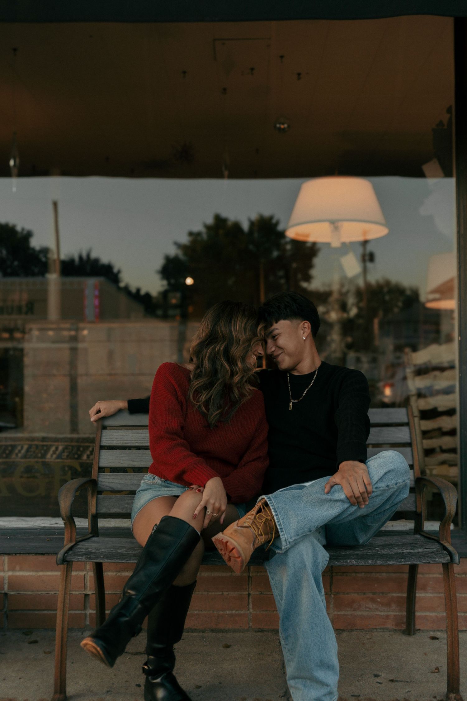 Romantic couple leaning in close on a bench outside a storefront at twilight, holding hands tenderly.