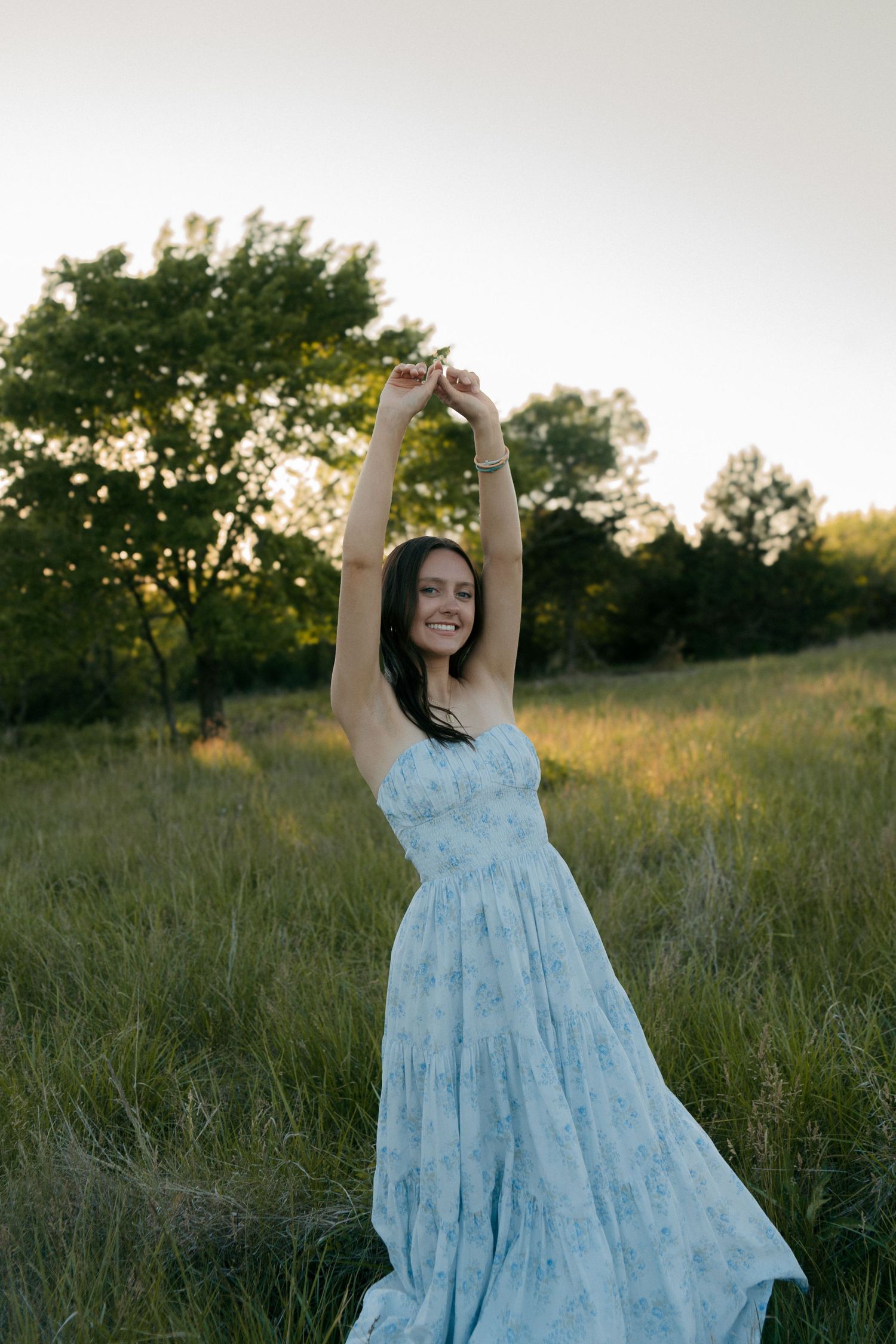 Young woman in light blue floral sundress dancing in green meadow at sunset with arms raised overhead.