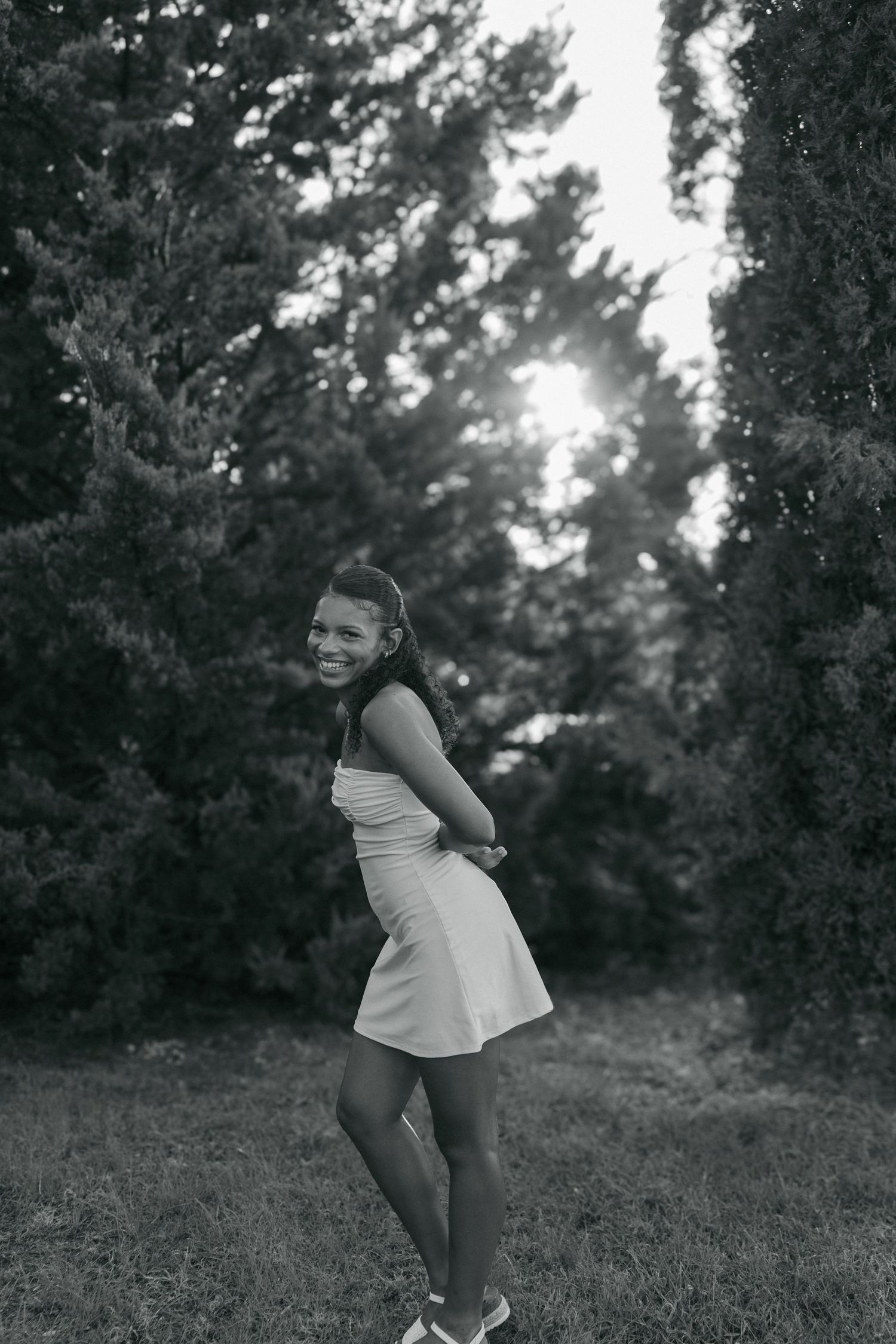 Smiling young woman in white dress posing playfully outdoors between cypress trees in black and white photo.