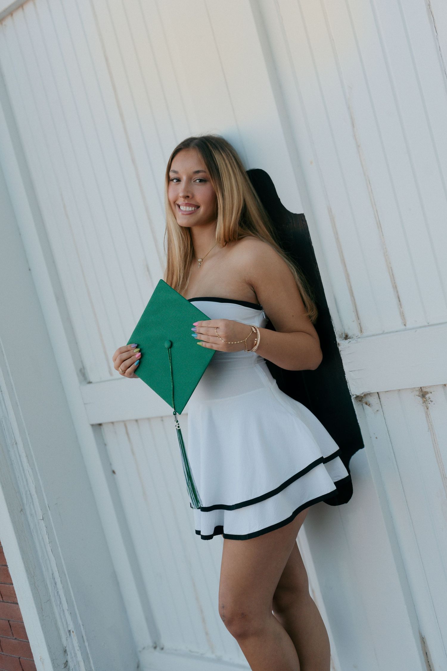 Smiling graduate in white dress holding green cap against white wooden wall.