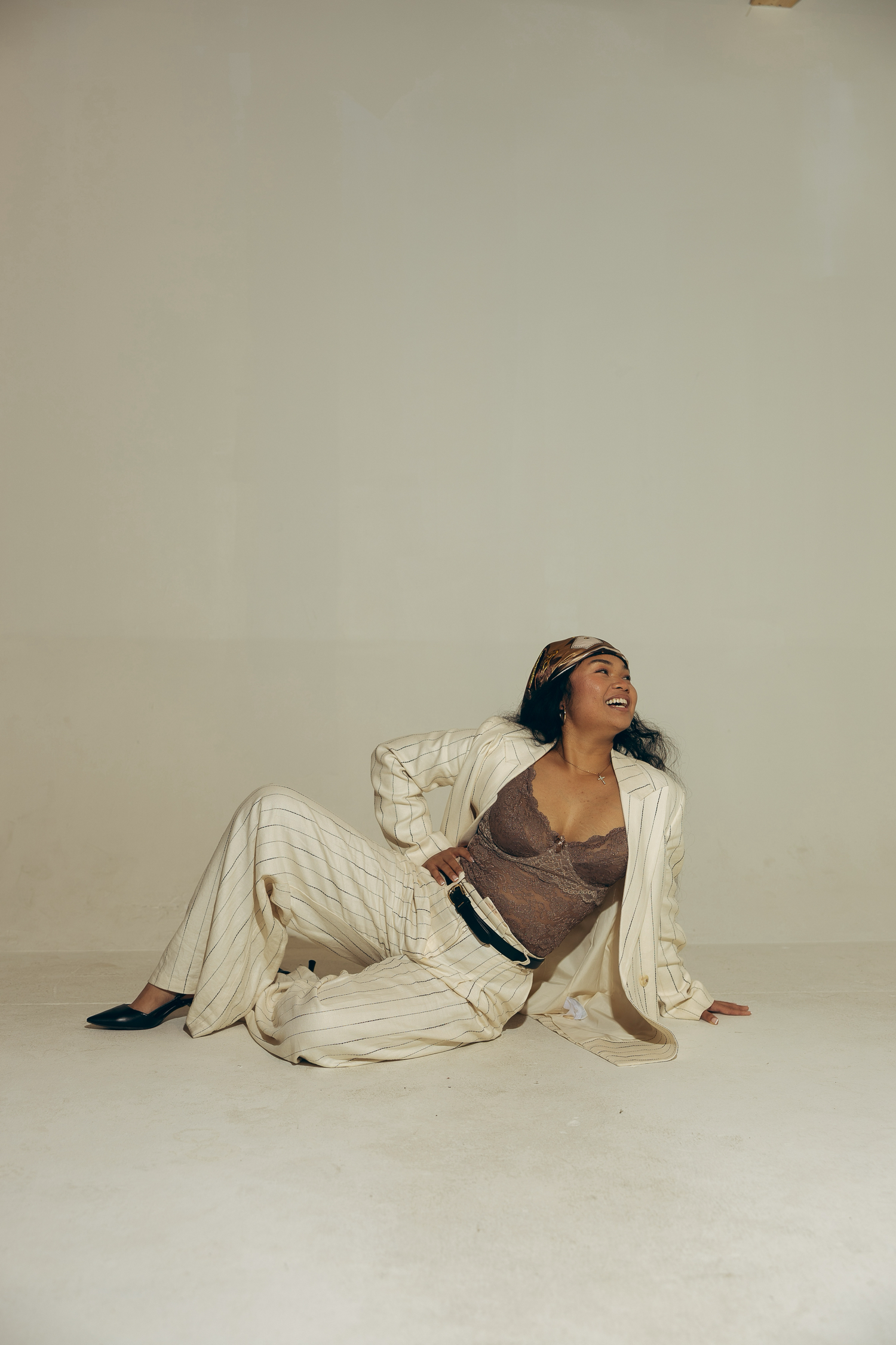Woman in white pinstripe suit sitting on floor, smiling upward, wearing brown top and headband in studio setting.