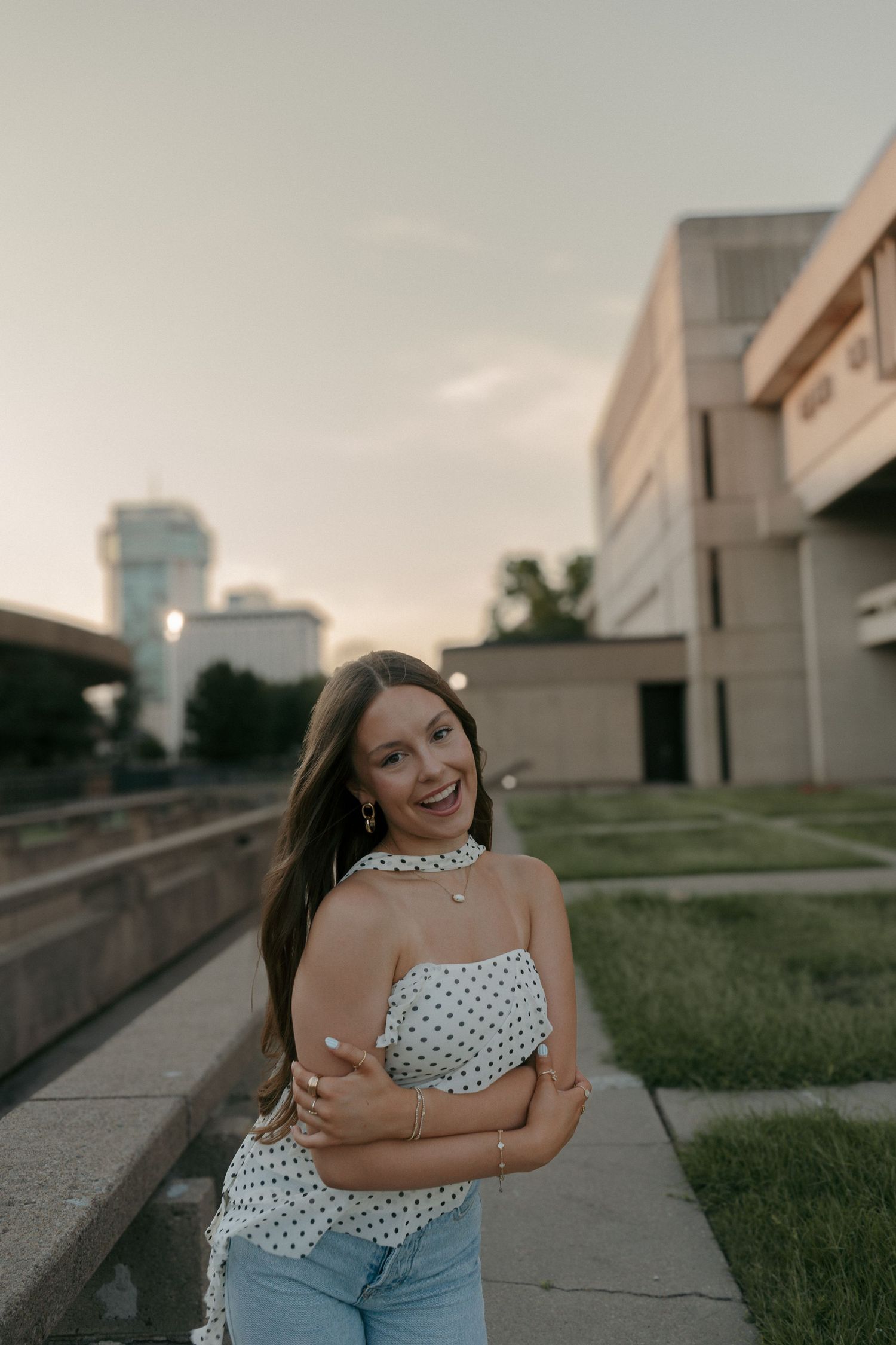 Laughing young woman in polka dot crop top and jeans poses on urban sidewalk at dusk.