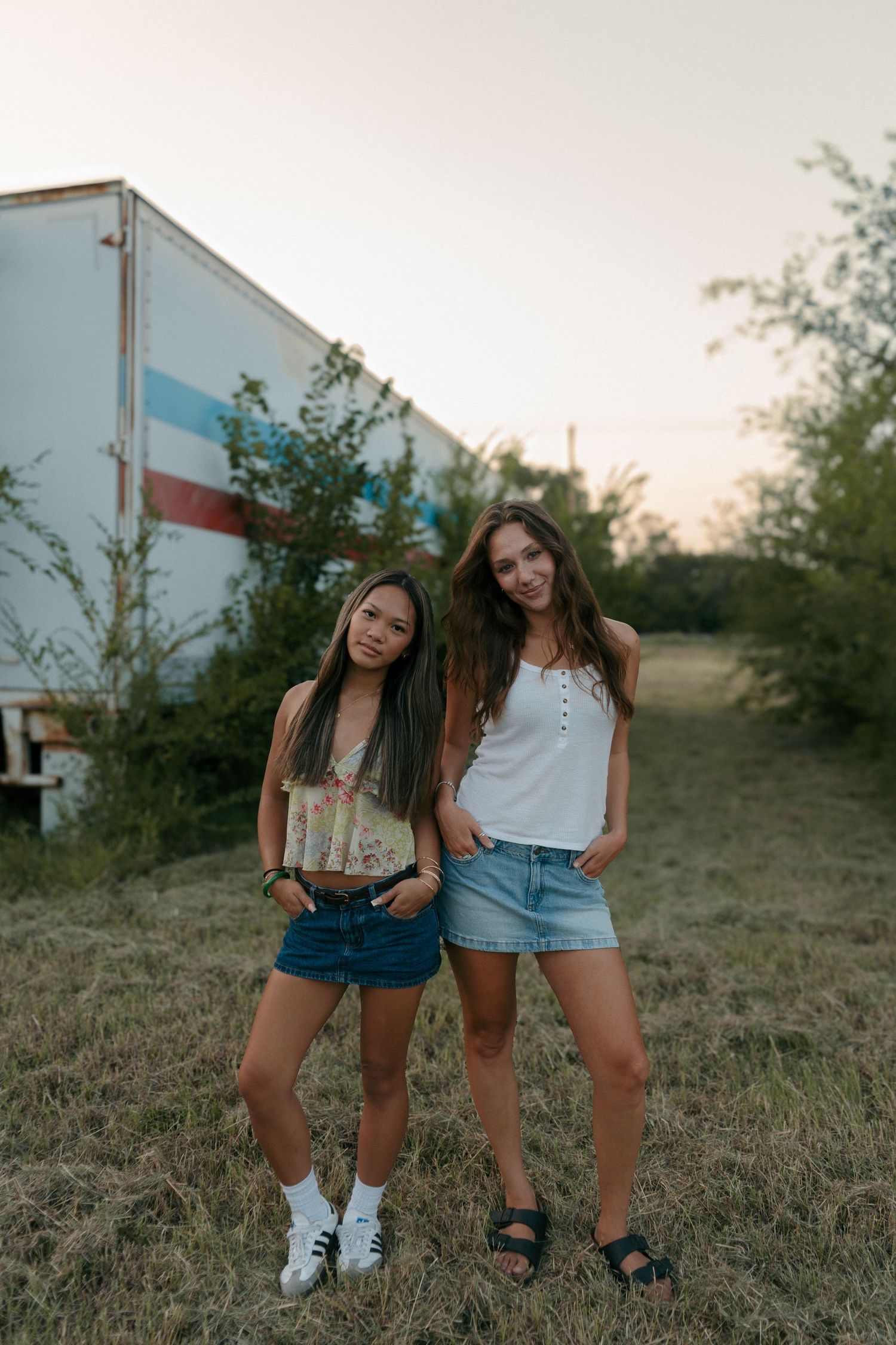 Two young women in denim skirts posing together in a field near a colorful trailer at dusk.