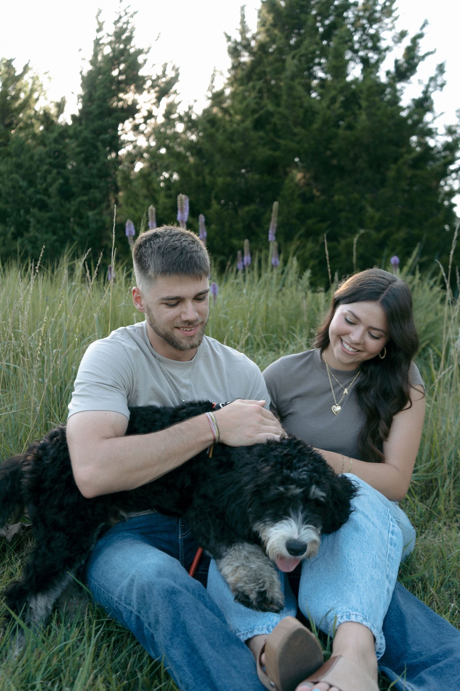 Happy couple sitting in green grass field petting their black and white Bernedoodle dog at dusk.