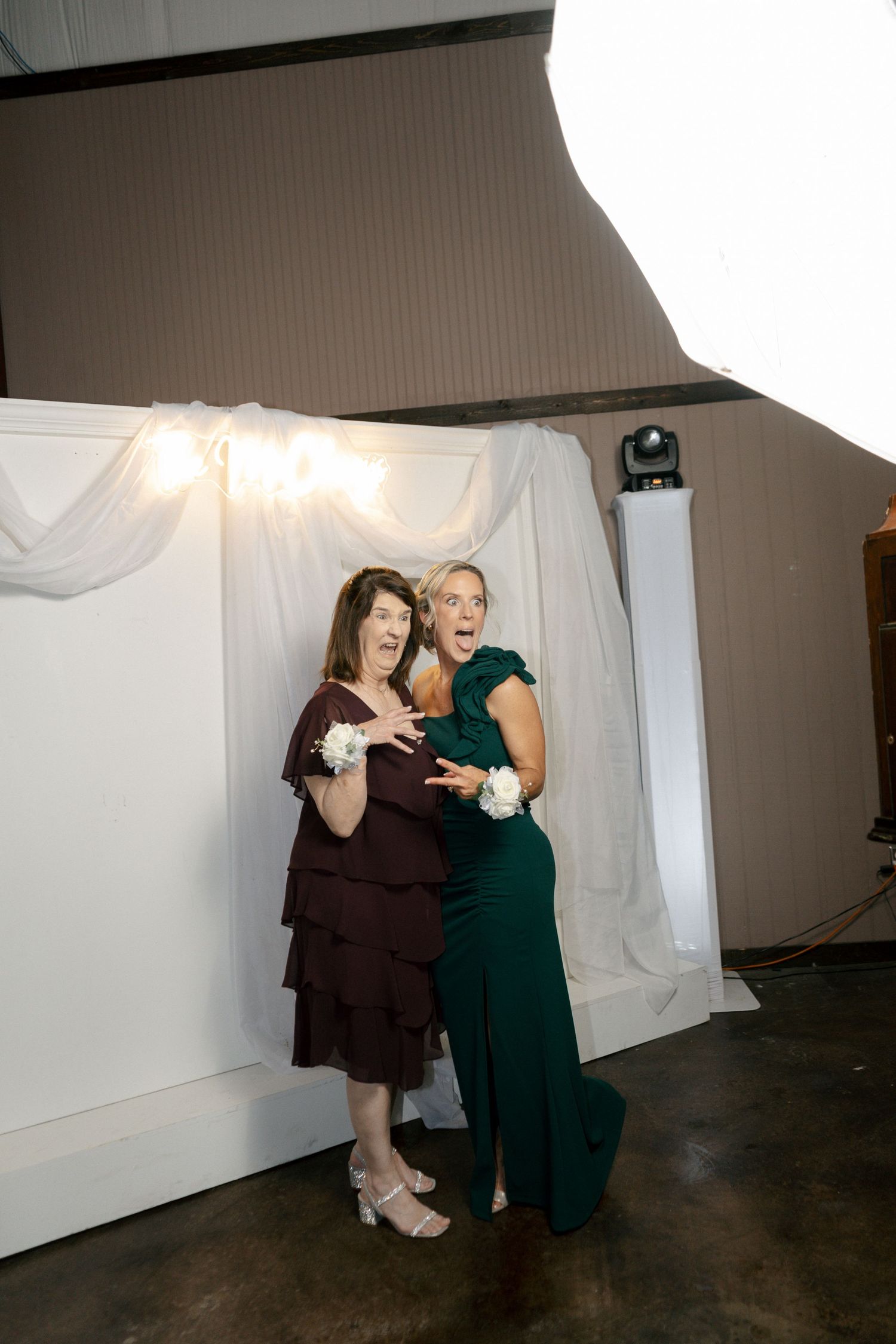 Two women in formal gowns pose playfully at a wedding photo booth, one in burgundy and one in emerald green.