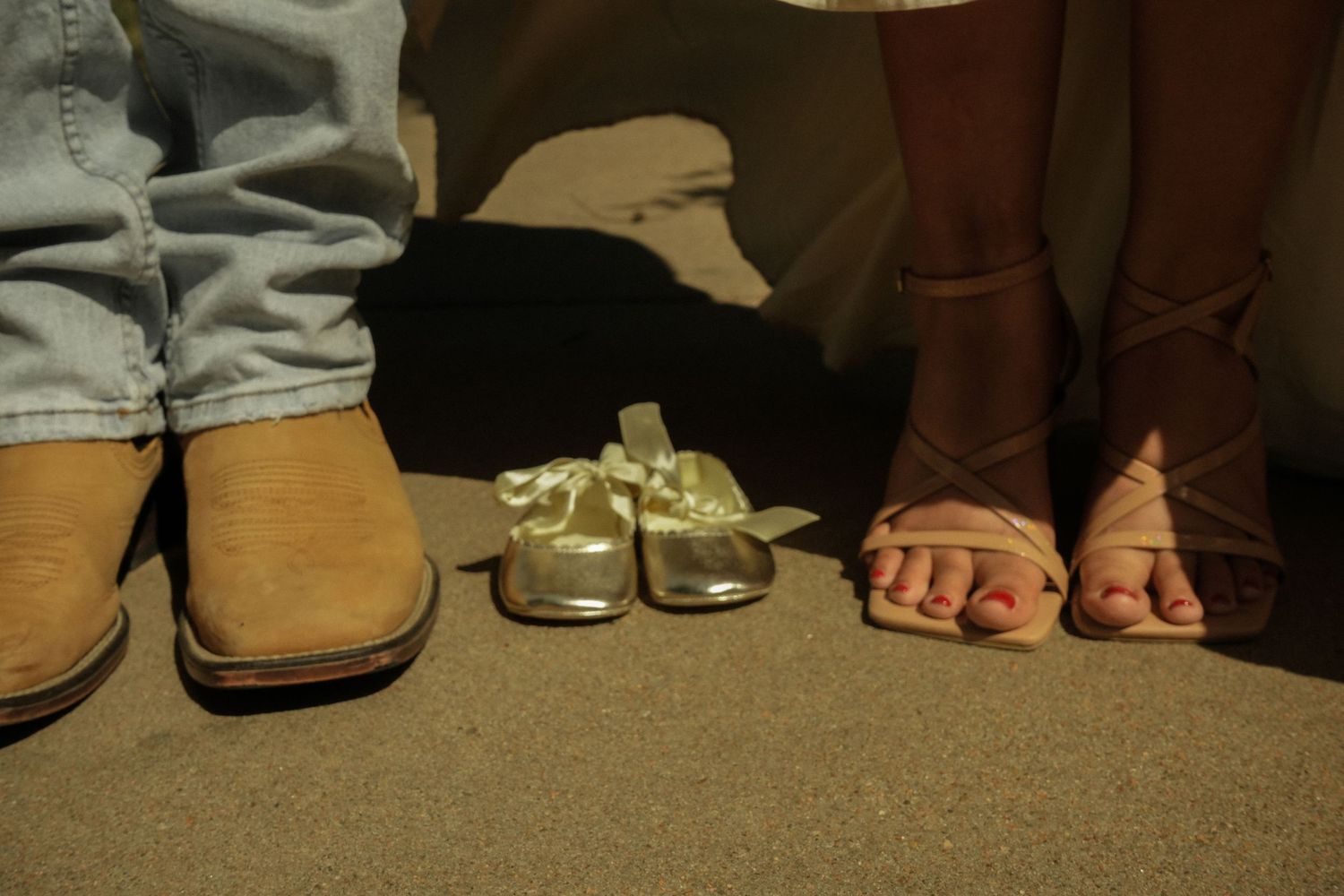 Couple's feet in cowboy boots and strappy heels with tiny gold baby shoes between them on sandy ground.