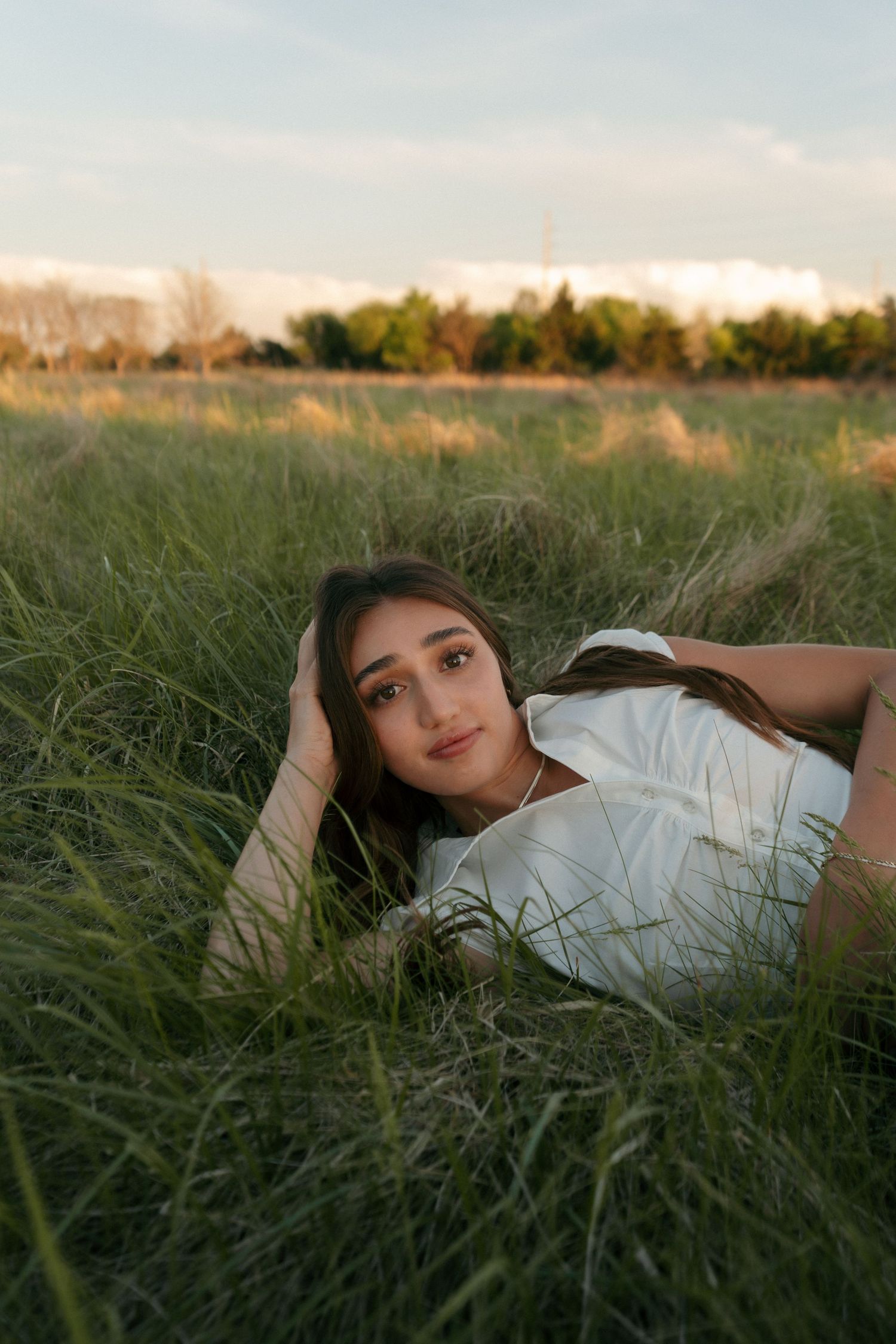 Young woman lying in tall green grass field gazing at camera during soft golden hour light.
