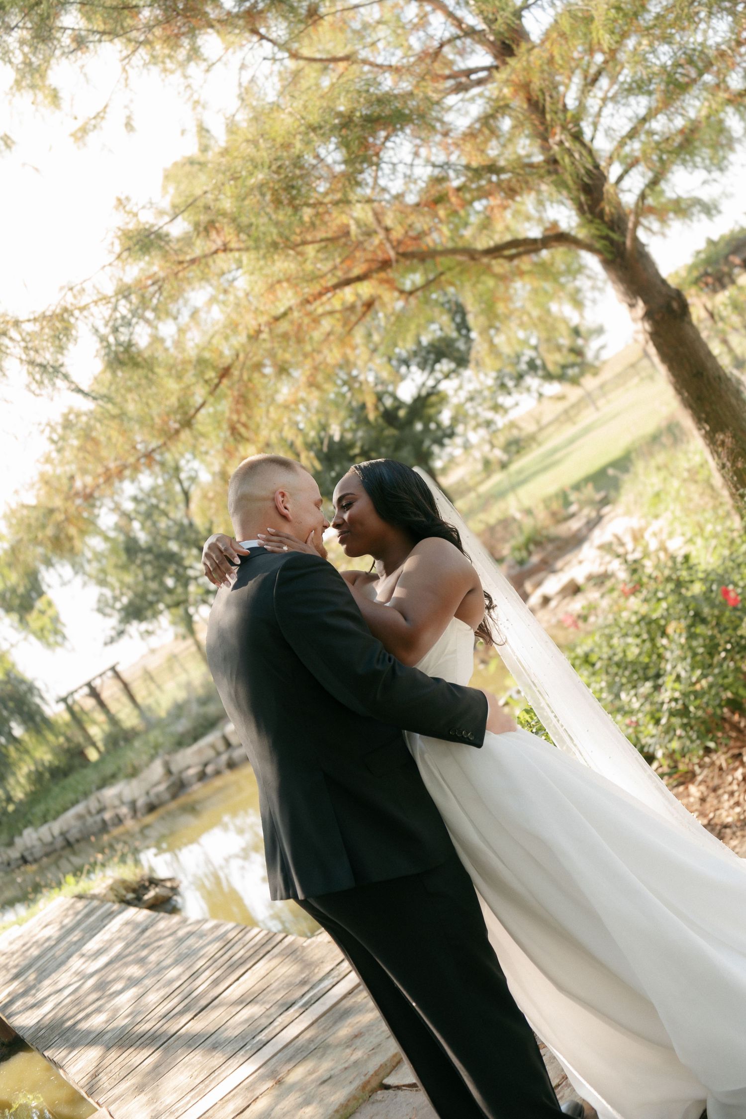 Bride in flowing veil and groom share romantic kiss near sunlit pond during outdoor wedding portraits.