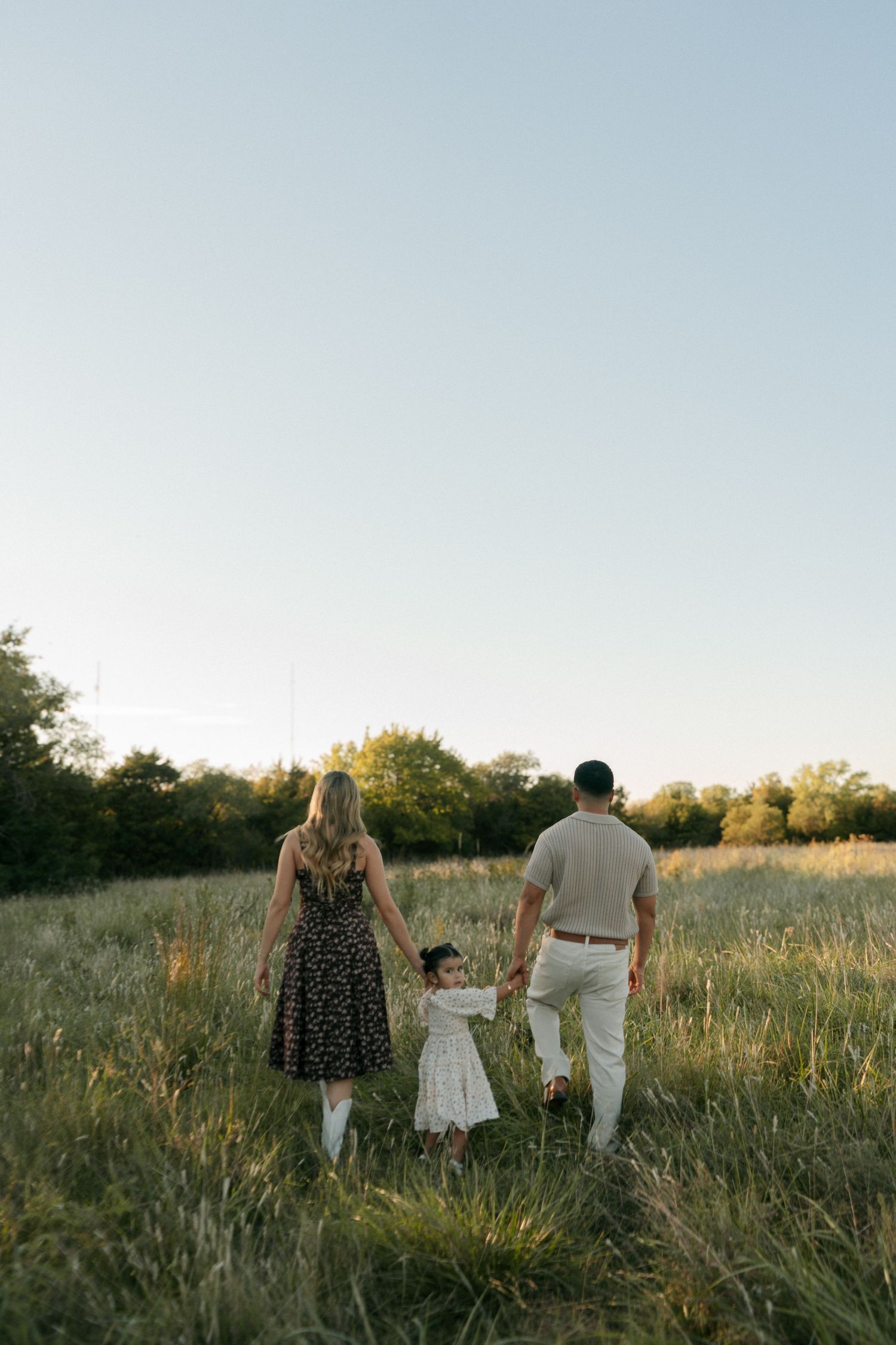 Family of three walking hand in hand through tall grass field at golden hour sunset.