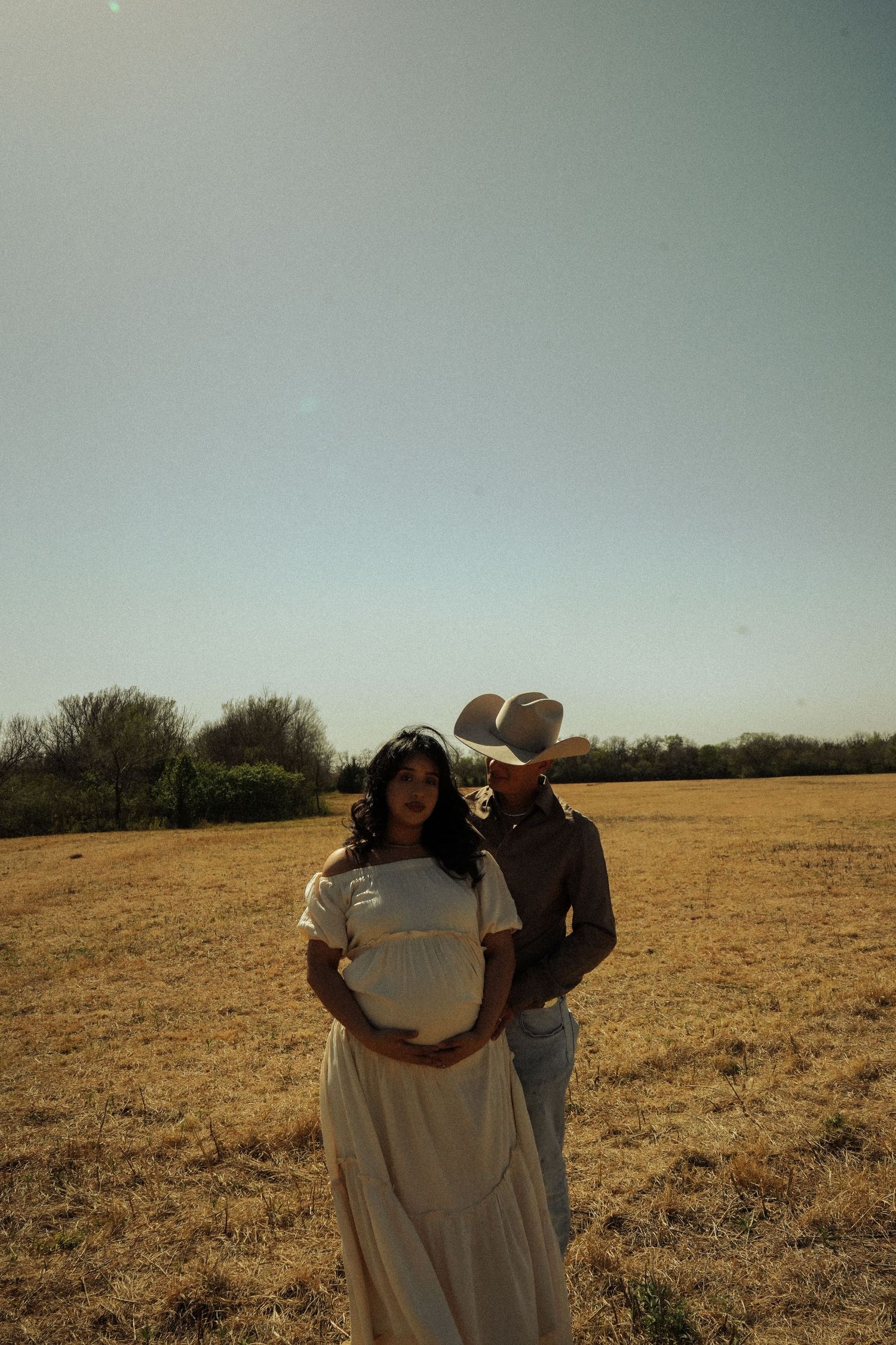 Pregnant woman in white dress with cowboy husband standing behind her in golden field under clear sky.
