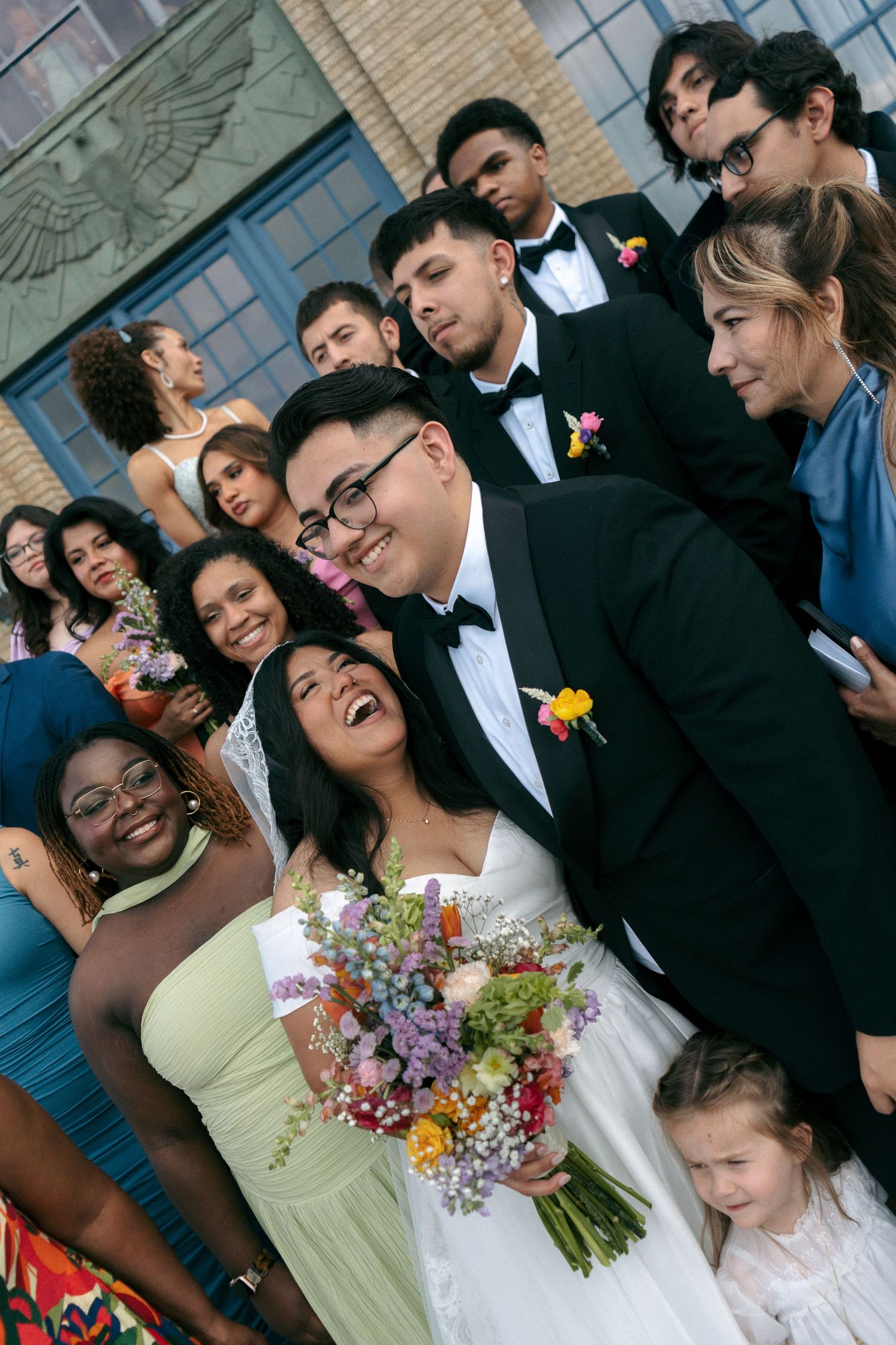 Joyful wedding couple poses with diverse group of guests outdoors, bride holding colorful bouquet in elegant attire.