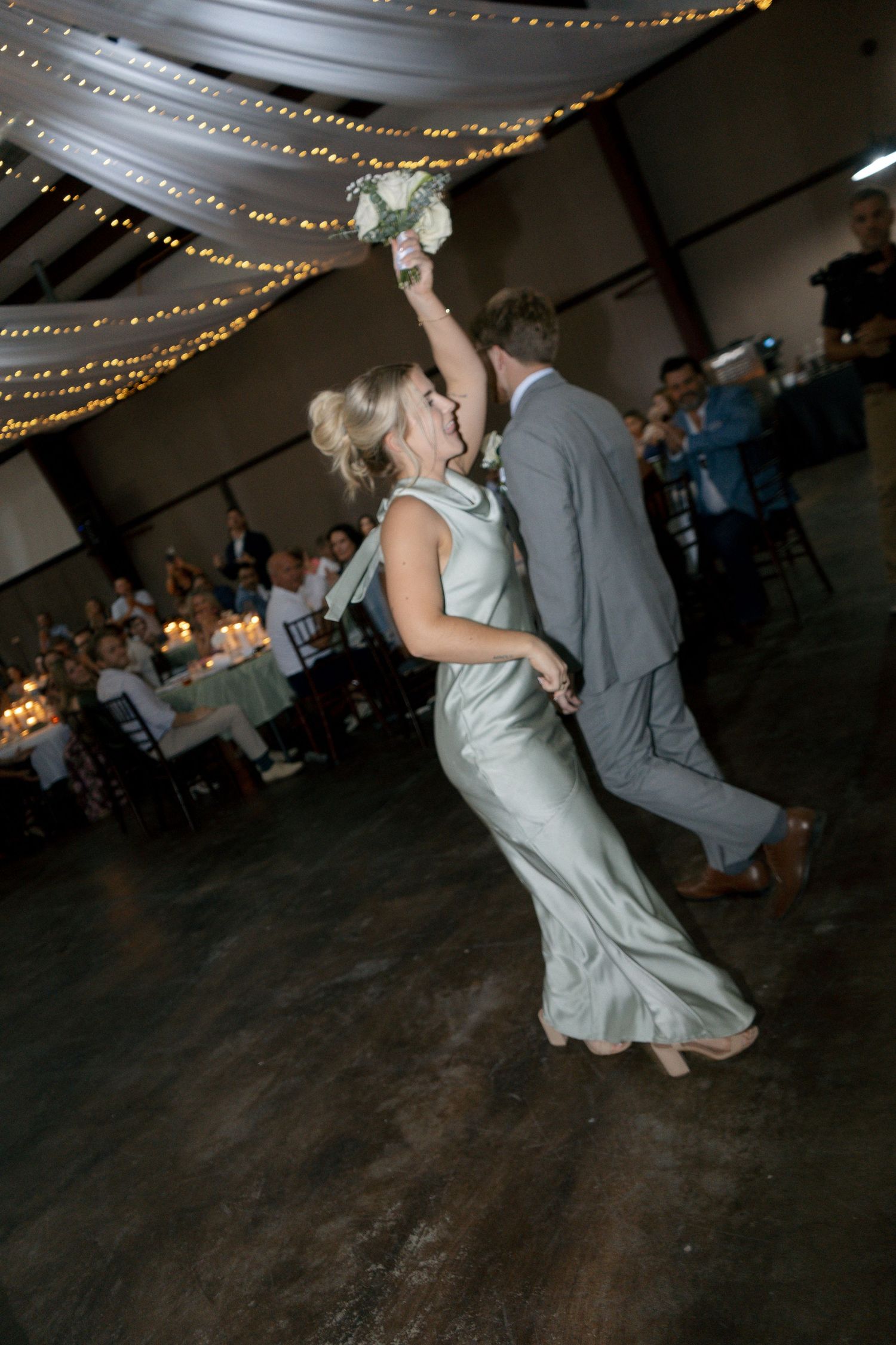 Bride in sage green gown raises bouquet while dancing with groom in gray suit at elegant wedding reception.