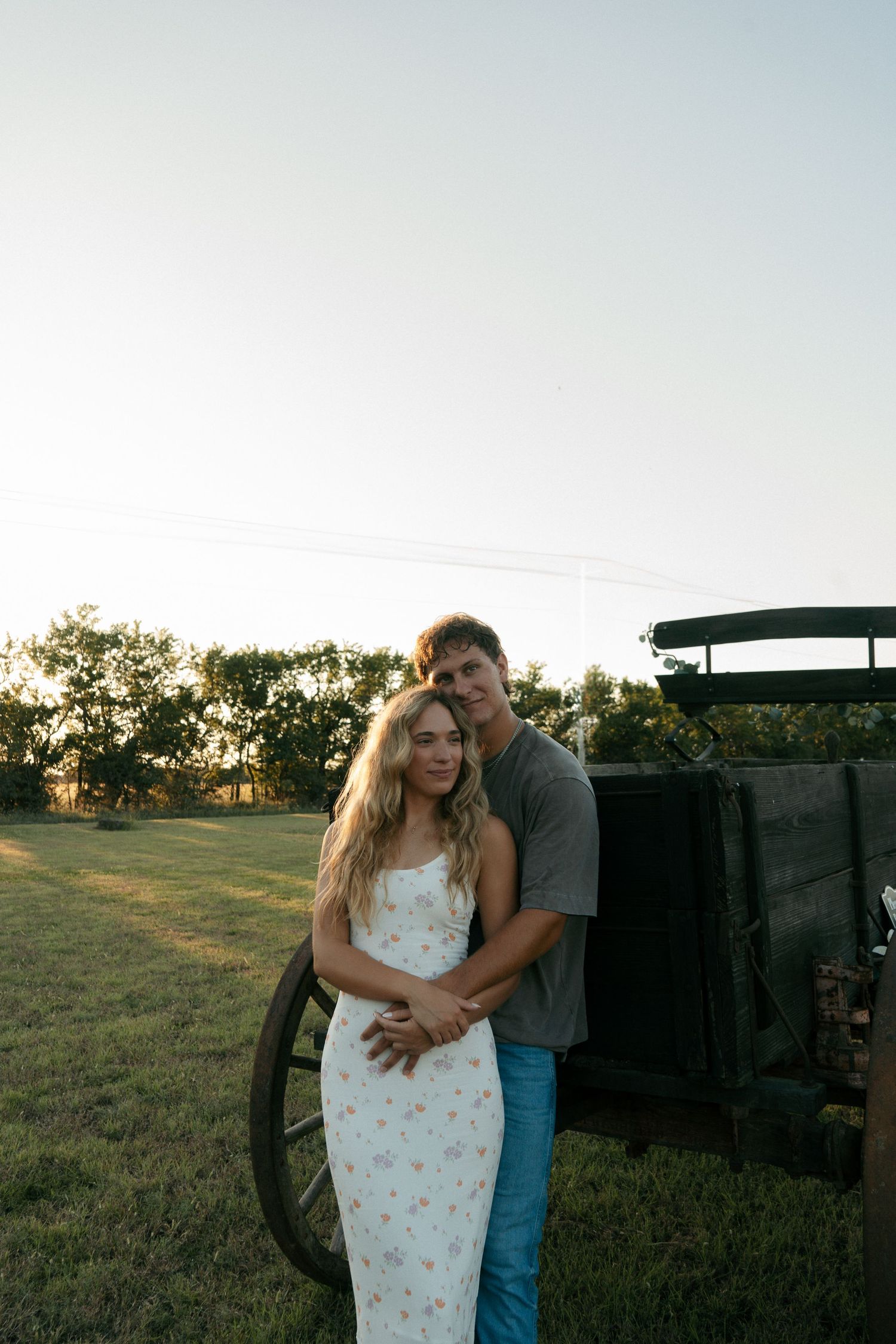 Young couple embracing near a rustic wooden wagon at golden hour on a green farm field.