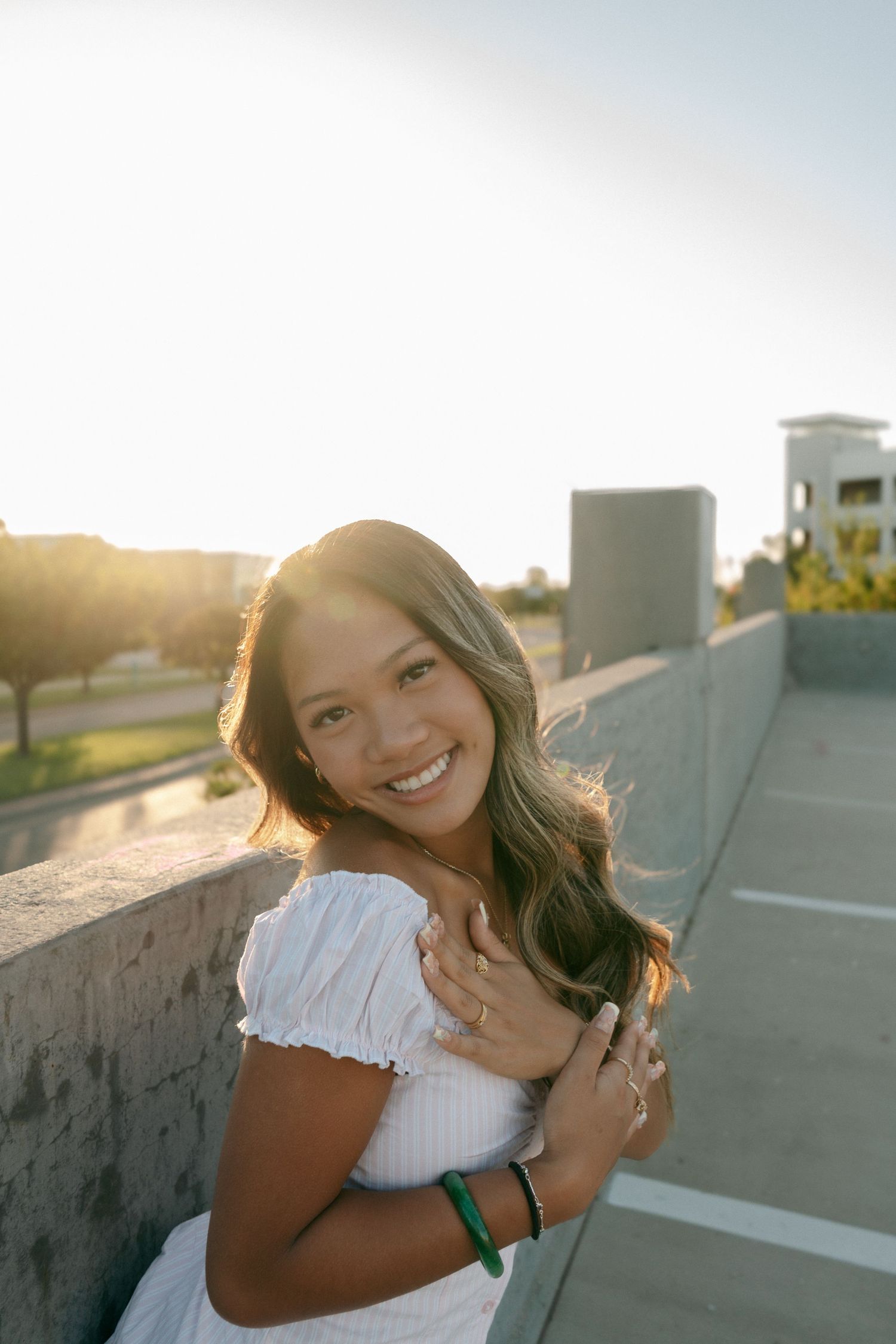 Smiling young woman in white off-shoulder top posing at sunset on rooftop parking structure.