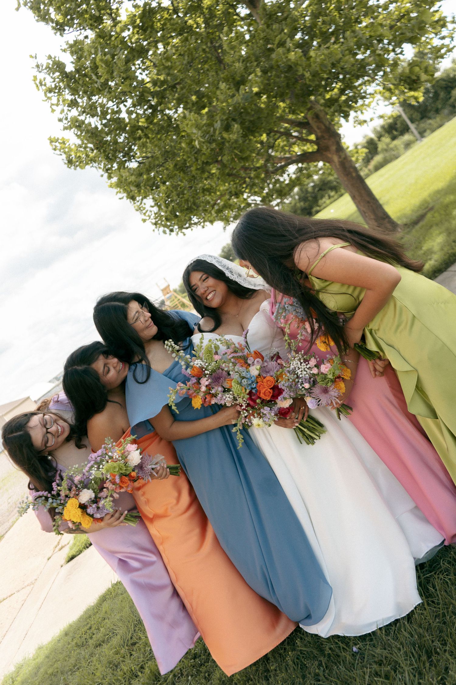 Bride in white gown with bridesmaids in colorful dresses holding floral bouquets outdoors near a tree and waterfront.