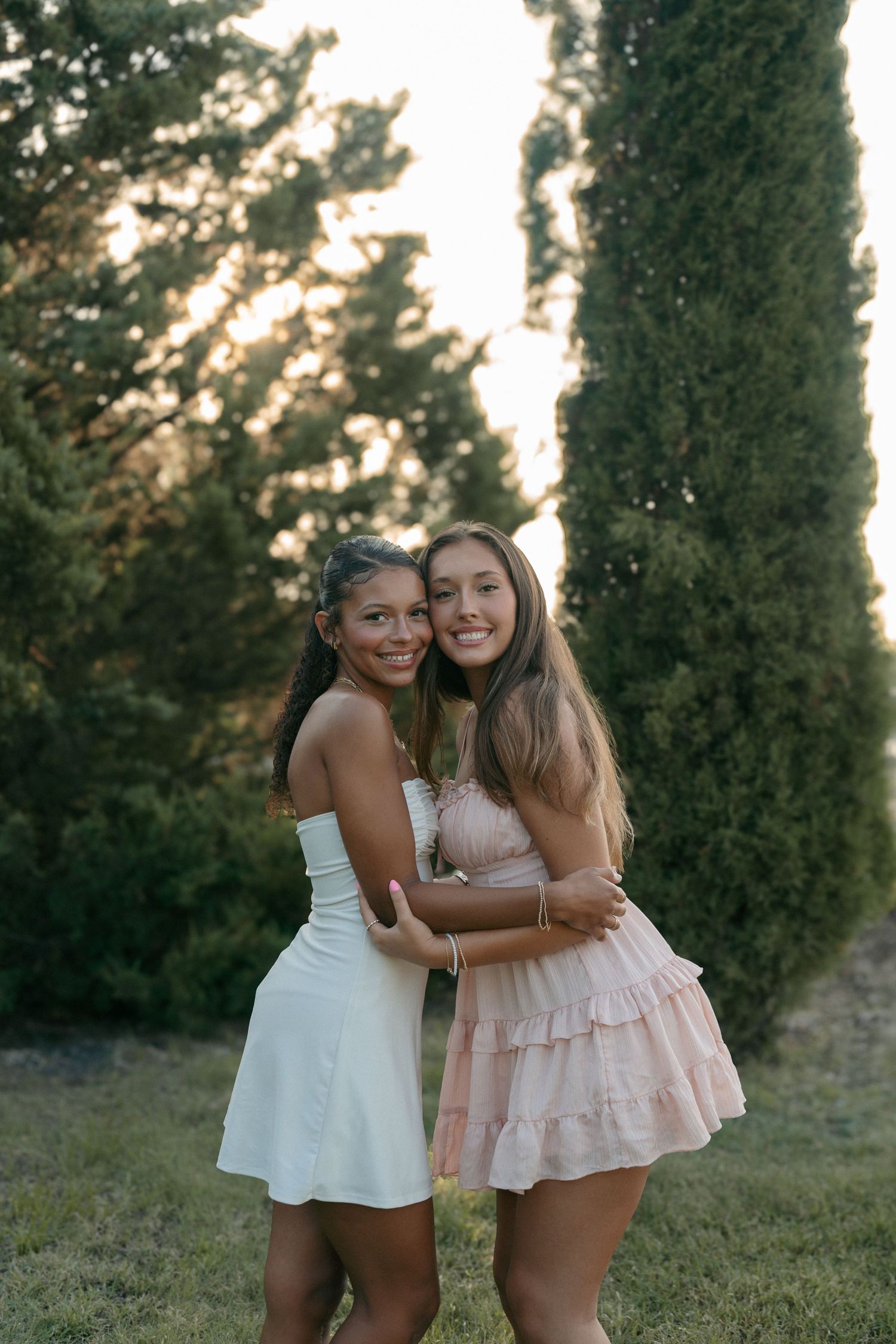 Two smiling young women in summer dresses hugging outdoors among tall cypress trees at golden hour.