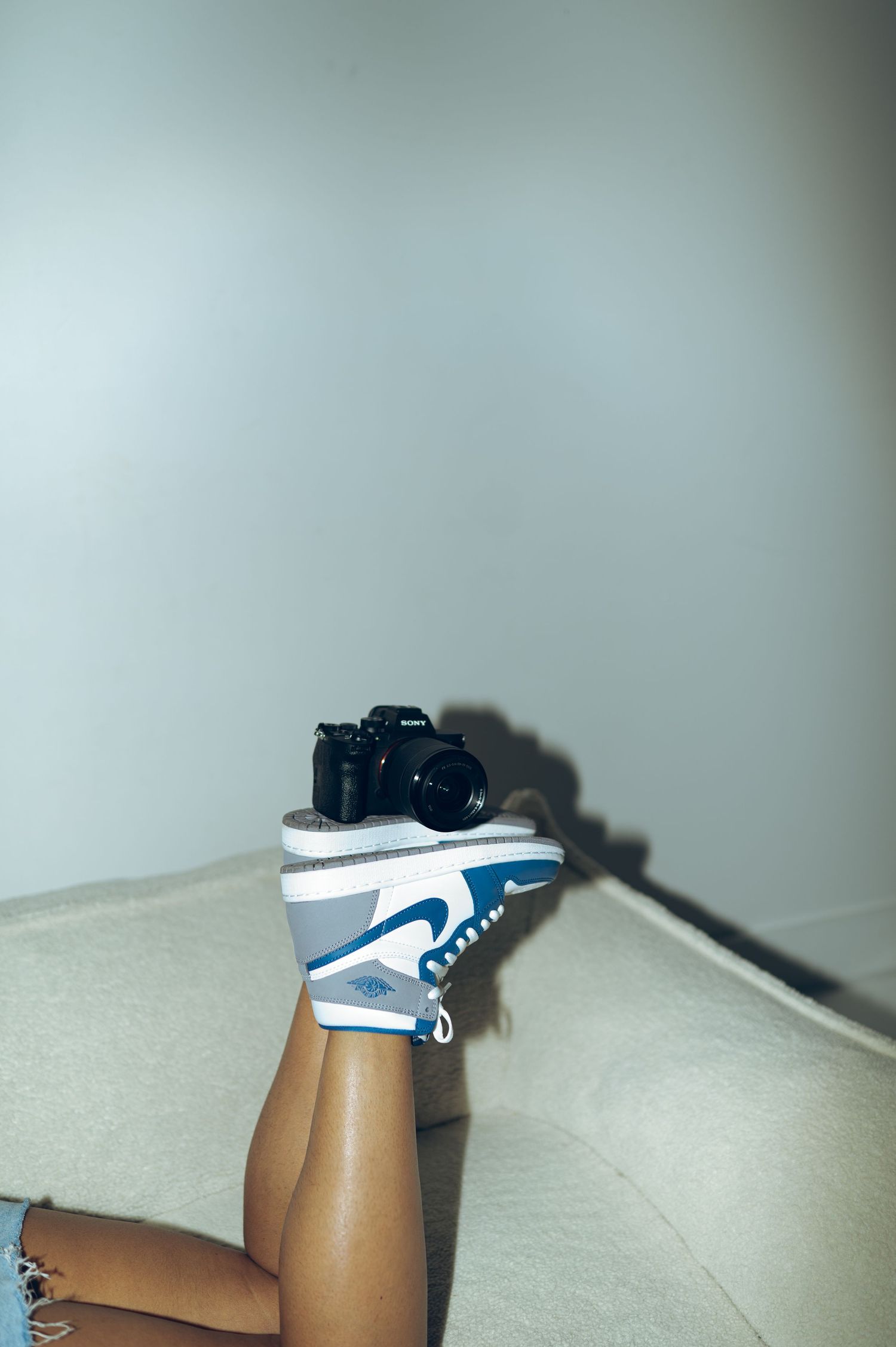 Legs raised on couch wearing blue and white Nike Air Jordan 1 sneakers with a camera balanced on top.