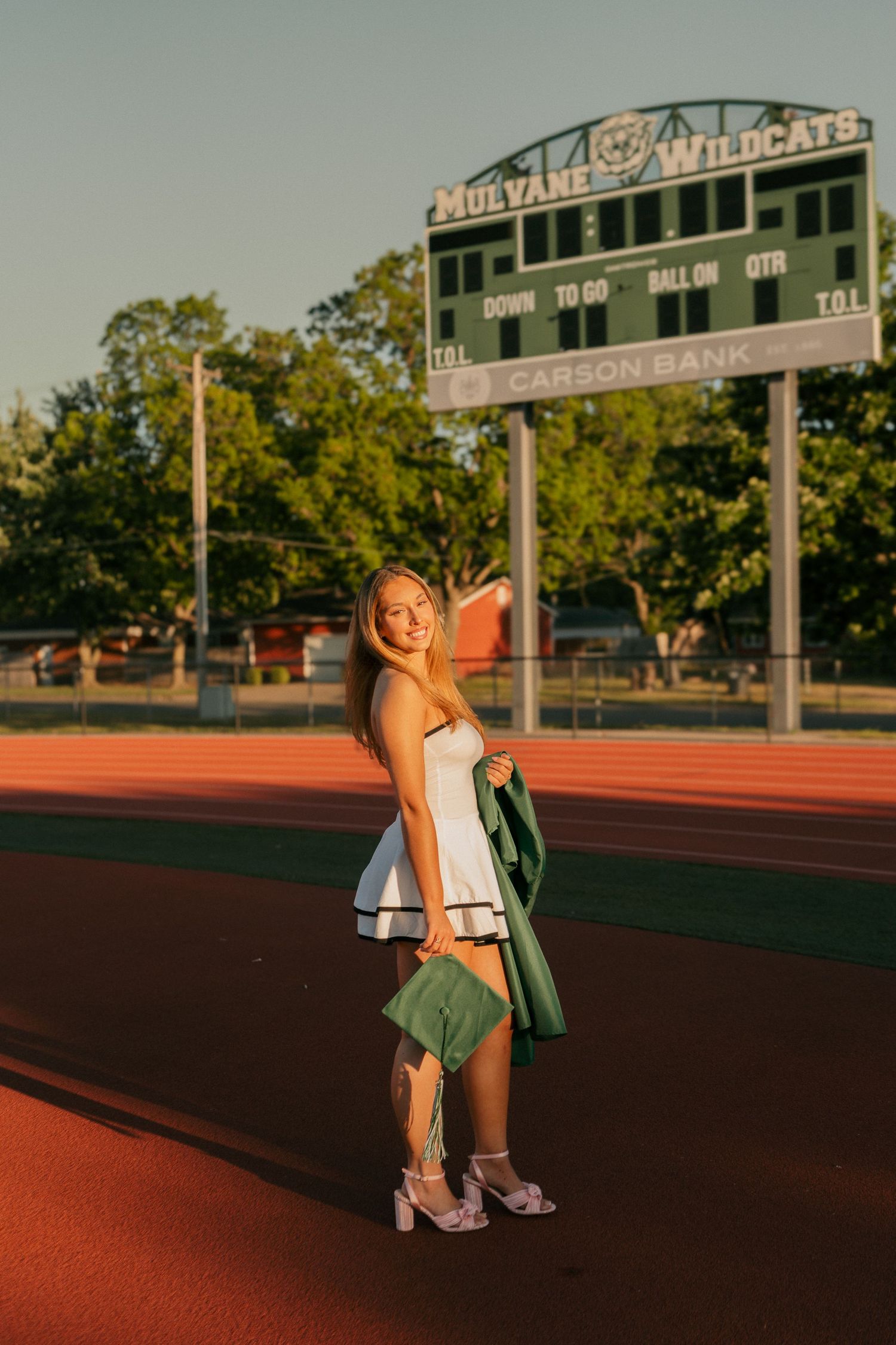 Graduate in white dress holding green cap near Mulvane Wildcats scoreboard at sunset.
