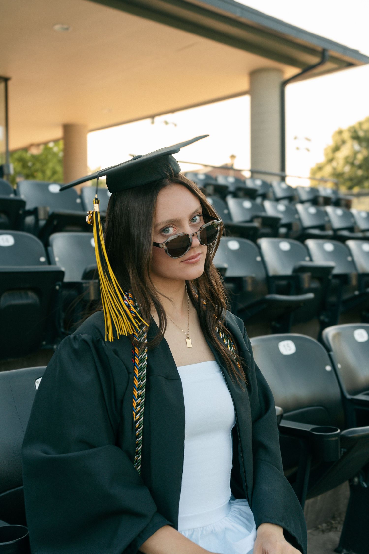 Graduate in black cap and gown wearing sunglasses seated in stadium bleachers at sunset.