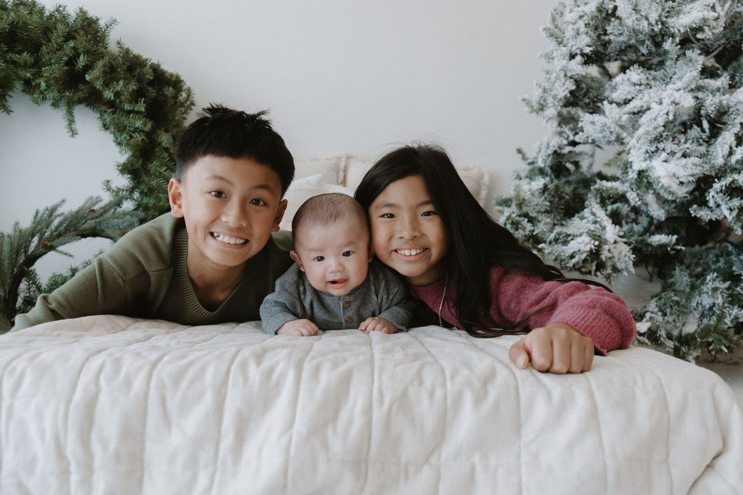 Three Asian siblings, including a baby, smile together on a white bed with Christmas decor behind them.