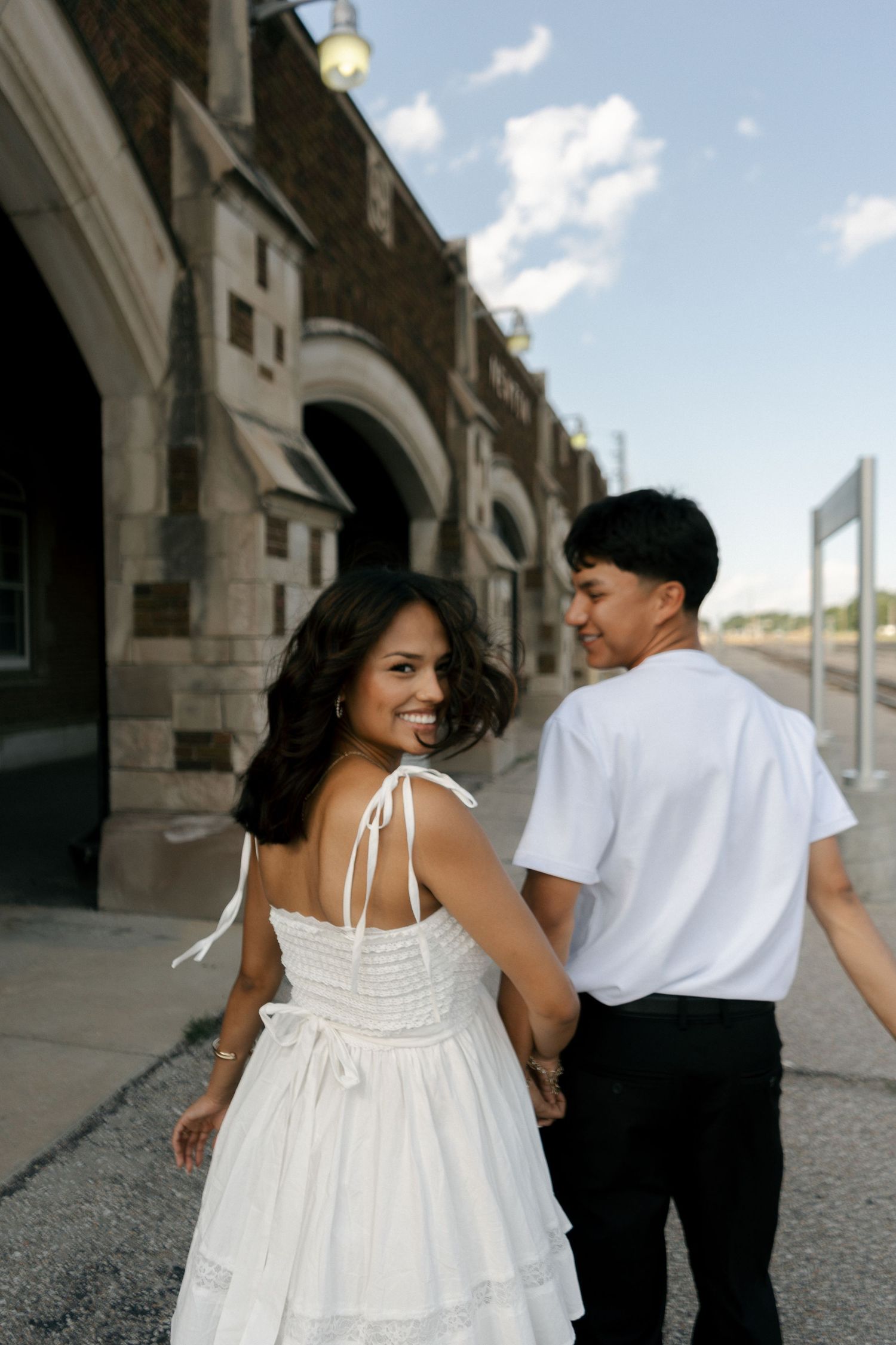 Happy couple holding hands and walking along a historic brick building, woman glancing back smiling.