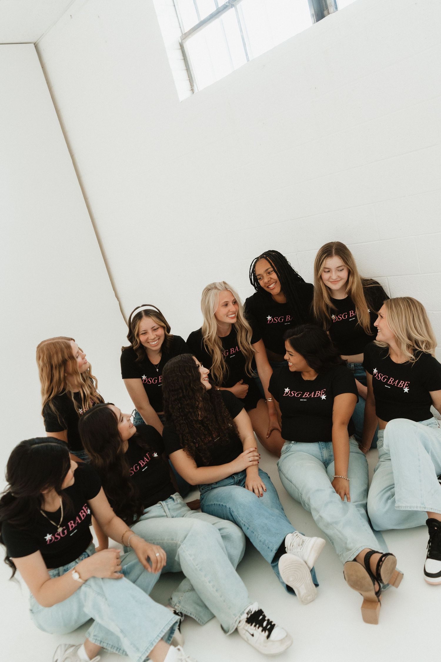 Group of women in matching black t-shirts sitting together laughing in a bright white studio setting.