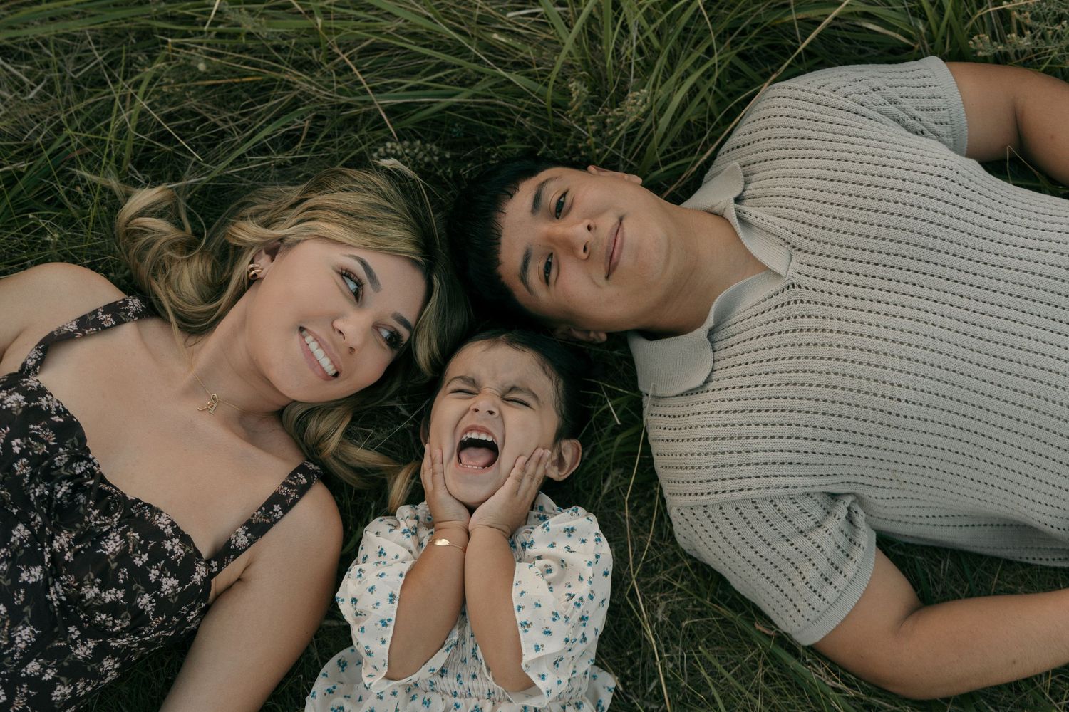 A joyful family of three lies in the grass laughing, with the young child in the middle making a funny face.