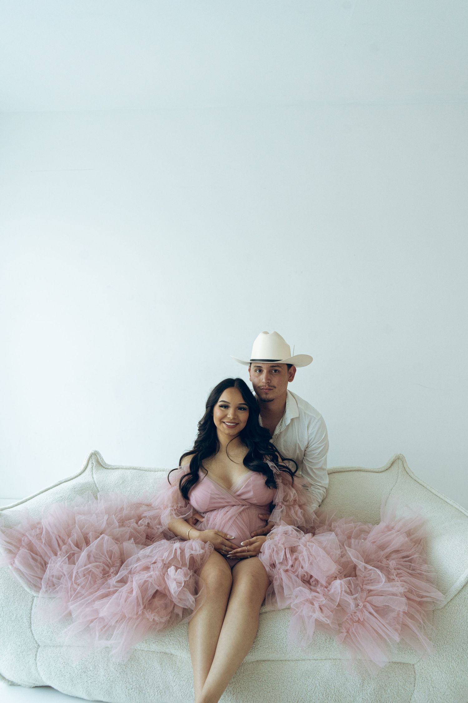 Couple smiles at camera during maternity photoshoot on white couch, woman in pink tulle gown and man in cowboy hat.