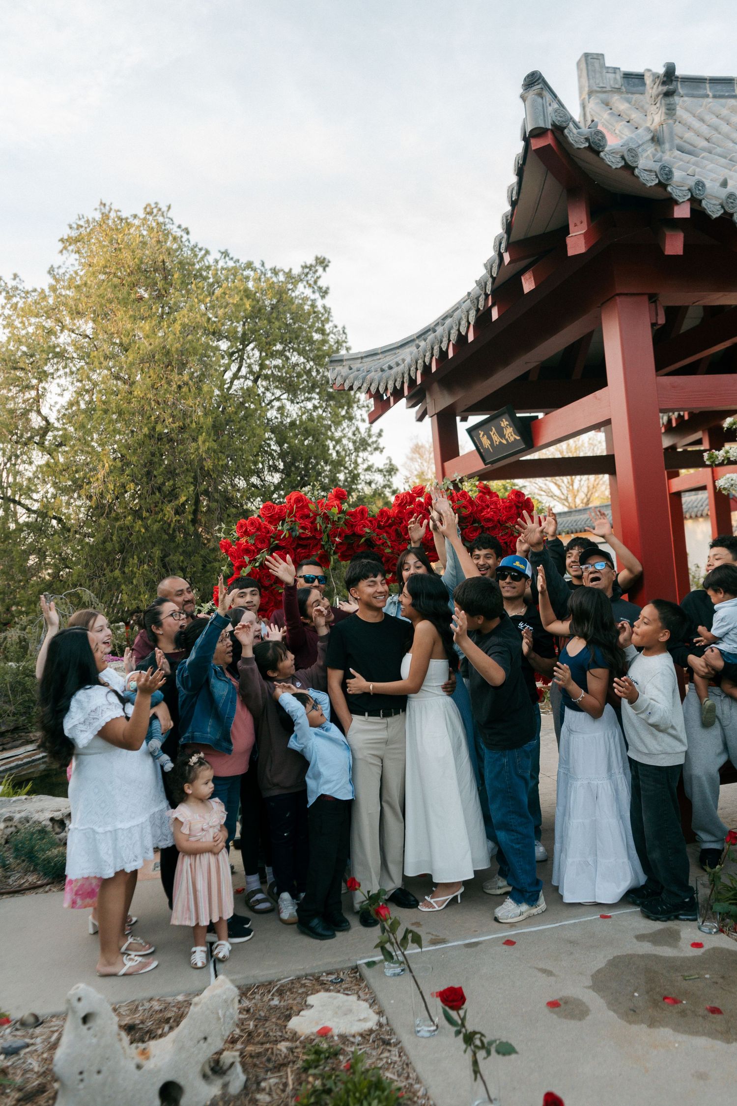 A large group of guests reaching up to catch a bouquet toss at an outdoor Asian garden wedding venue.