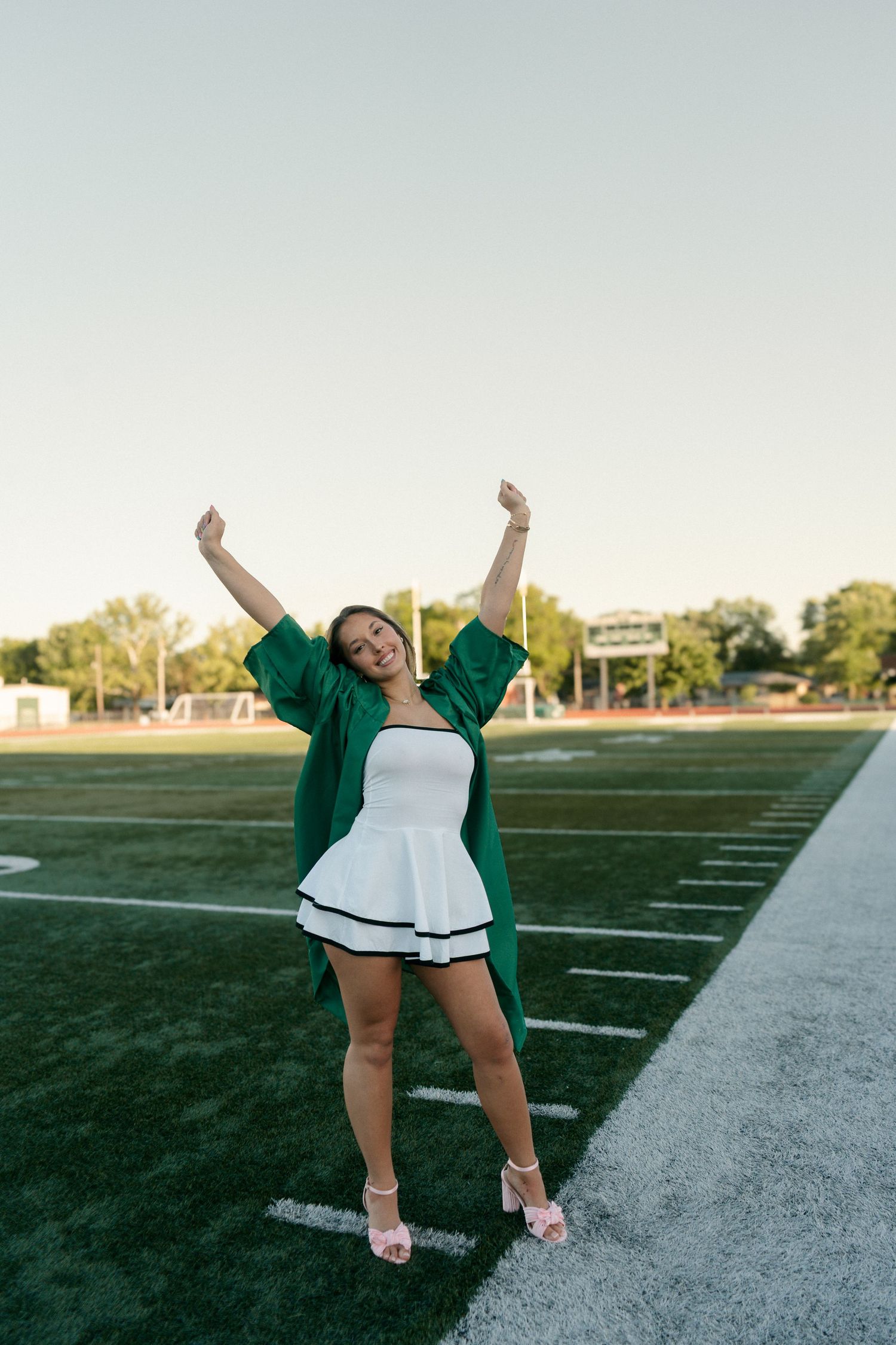 Graduate celebrating in green cap and gown on football field with arms raised.