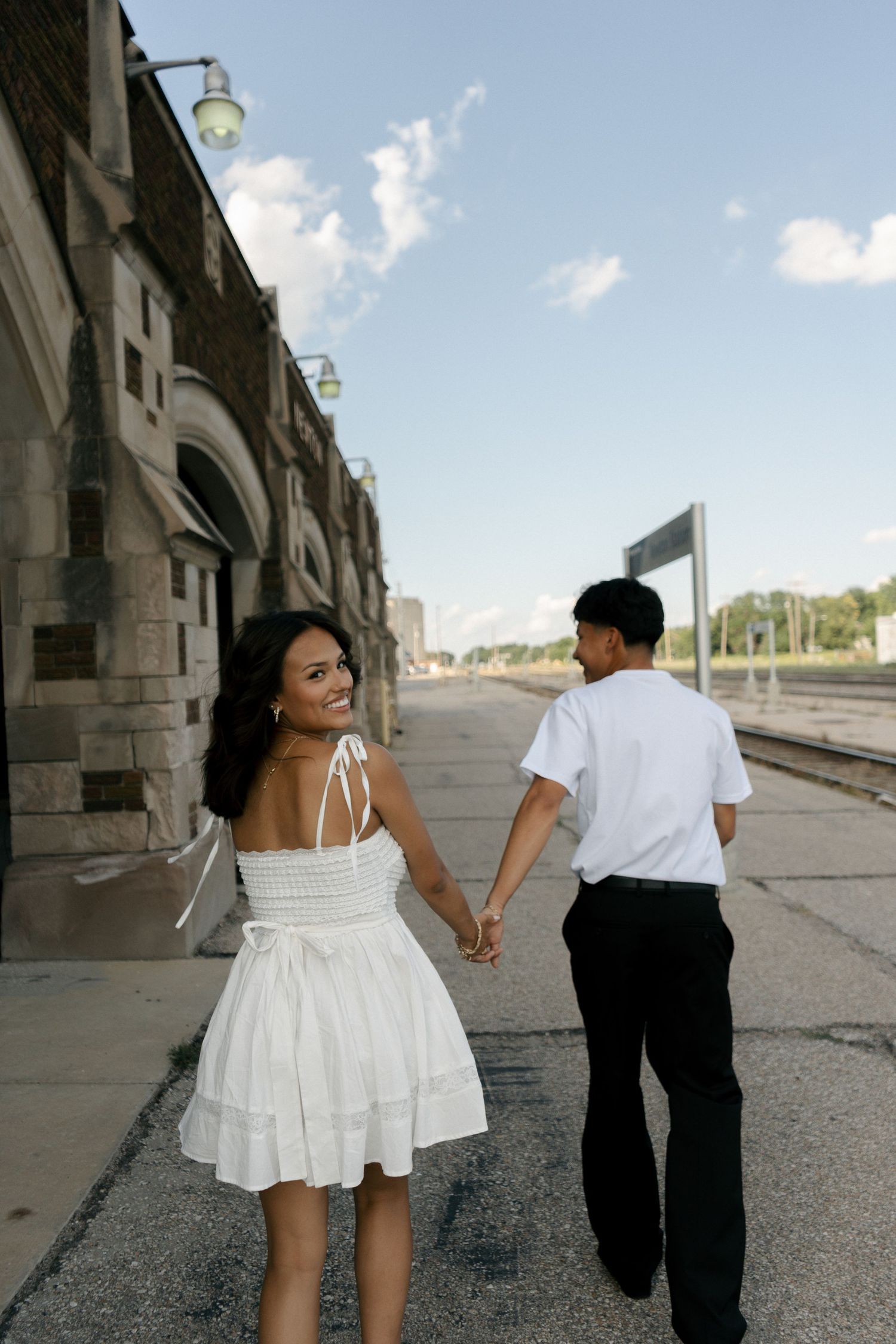 Smiling woman in white dress holds hands with man in white shirt walking along a train platform.